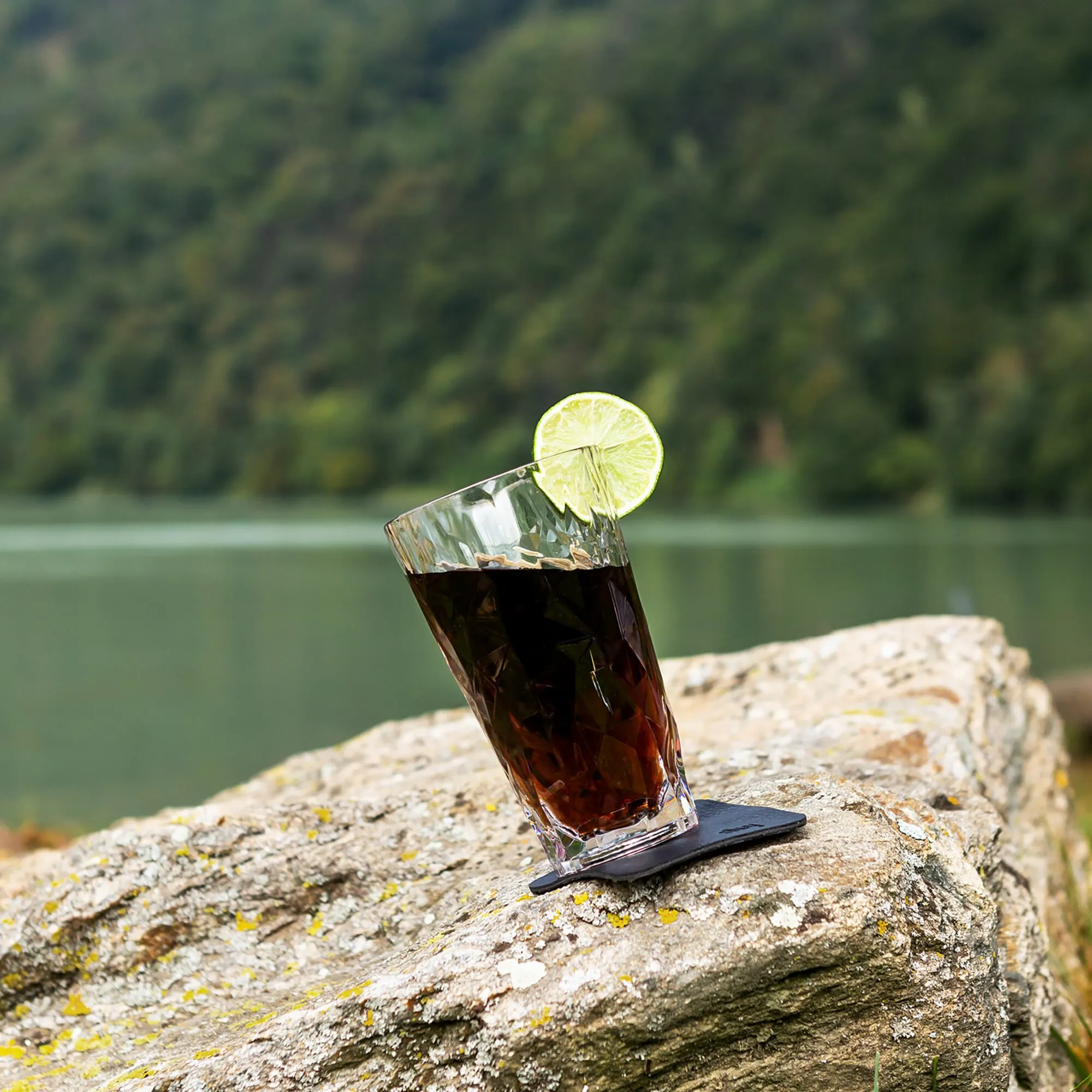 Facettiertes dunkles Longdrinkglas aus Kunststoff mit Limettenscheibe auf einem schwarzen Magnetuntersetzer auf einem Felsen