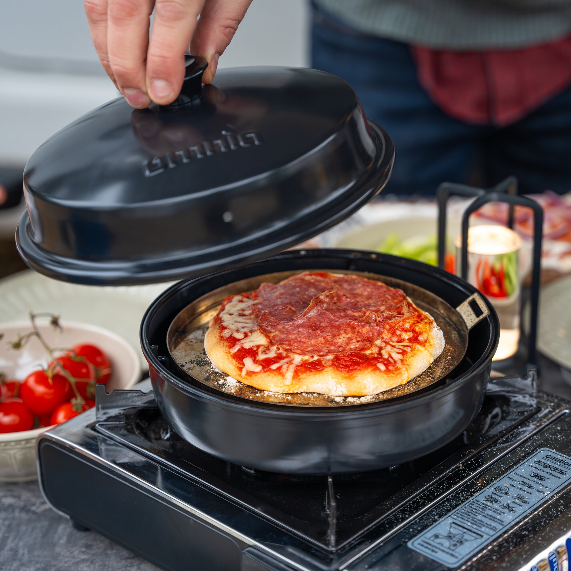Hand hebt den Deckel eines schwarzen runden Stovetop-Pizzaofen; darunter Pizza mit Käse und Tomatensauce, Tomaten in einer Schale