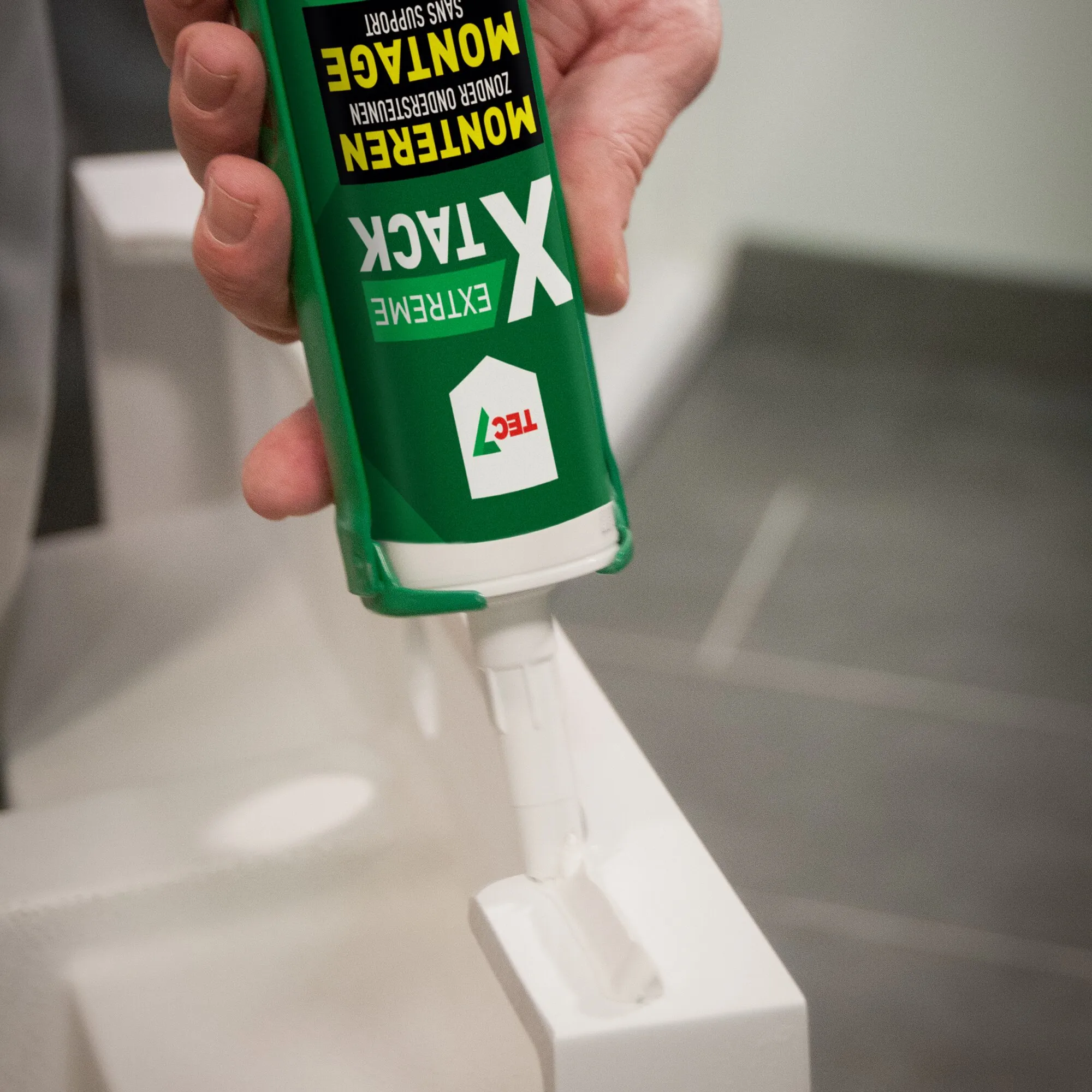Grüne Kartusche Montagekleber XTACK EXTREME mit weißer Kleberaupe auf einer weißen Leiste, von einer Hand gehalten