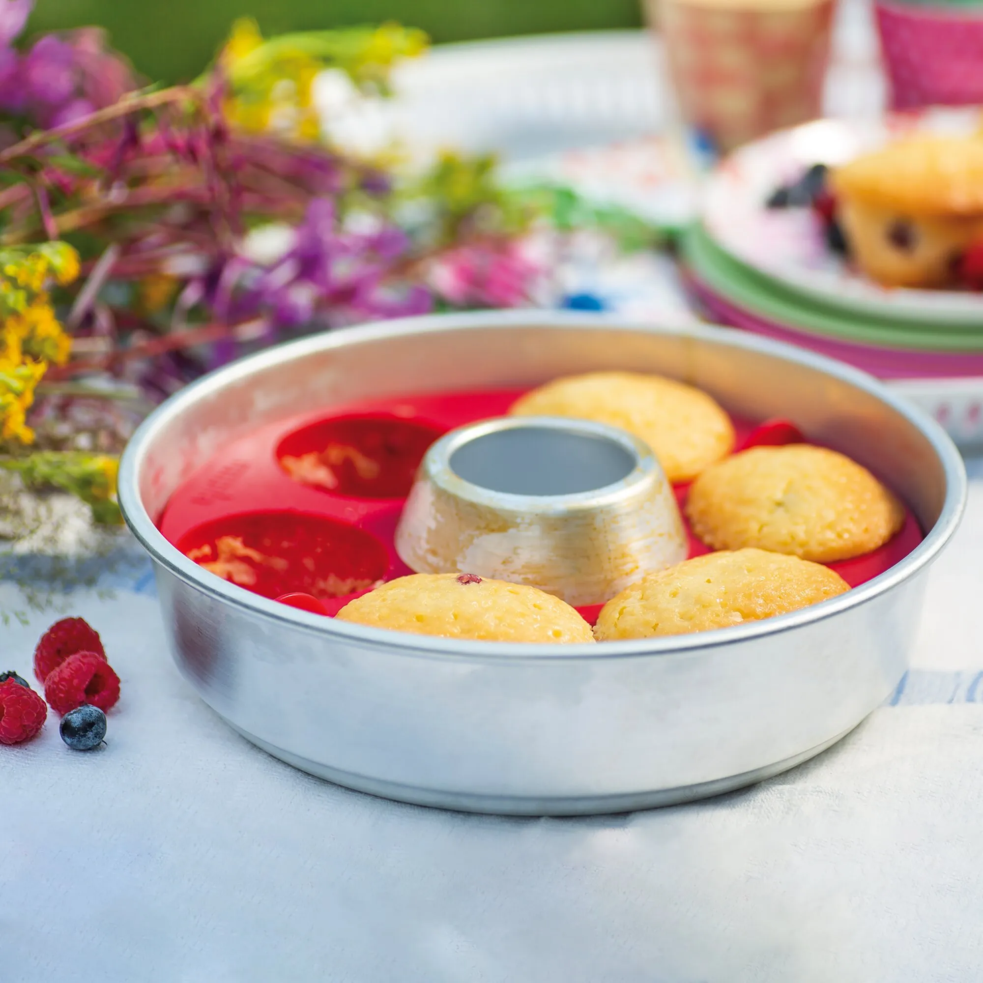 Muffin-Tab aus Metall mit sieben goldbraunen Muffins, roten Förmchen und zentraler Öffnung auf einem gedeckten Tisch