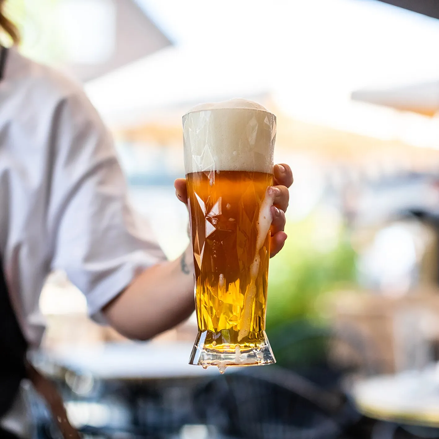 Person hält ein transparentes, facettiertes Weißbierglas CLUB mit goldenem Bier und üppiger Schaumkrone in der Hand