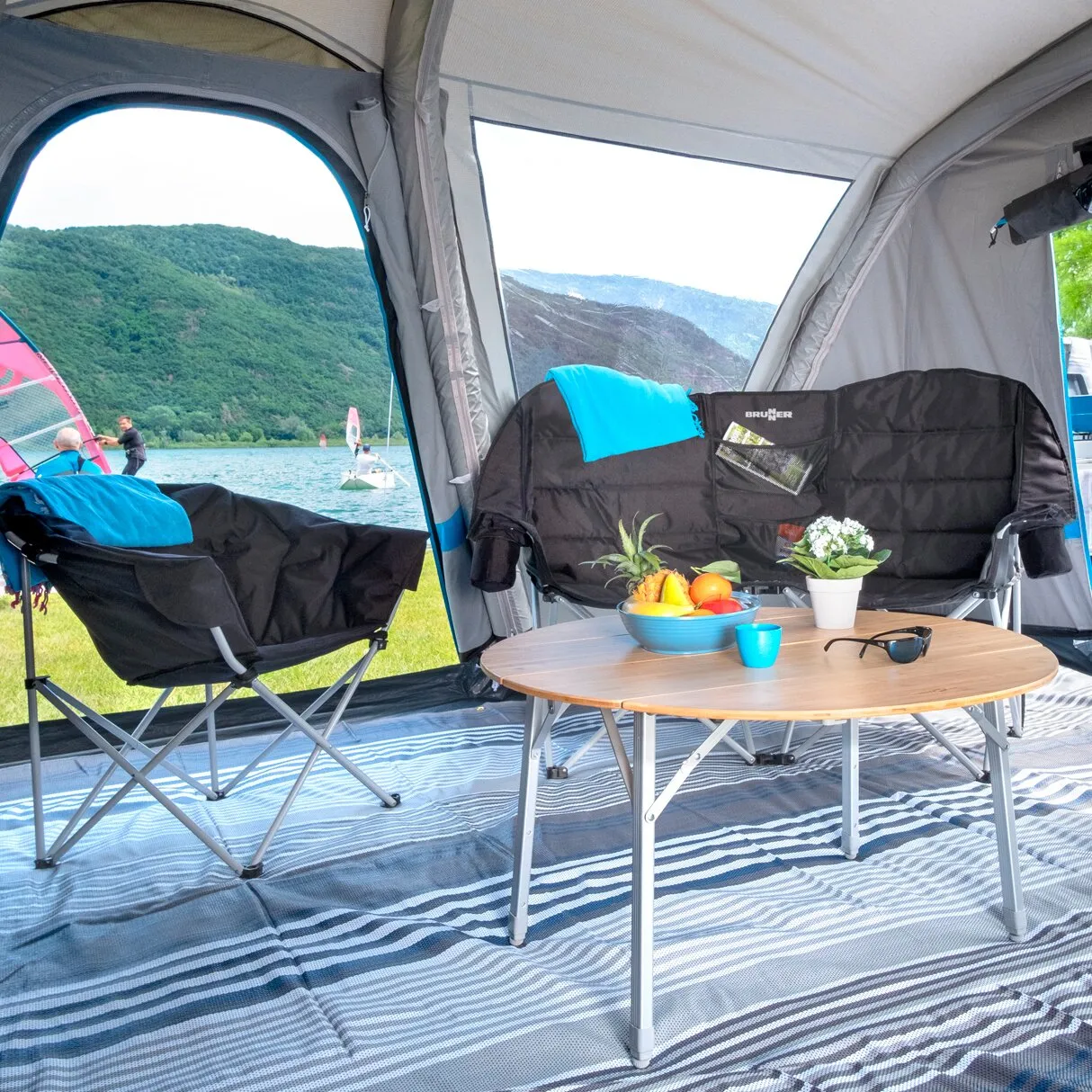 Schwarzes Faltsofa Rendez-Vous mit Armlehnen im Campingzelt, davor ein Klapptisch mit Obst und Getränken am Seeufer