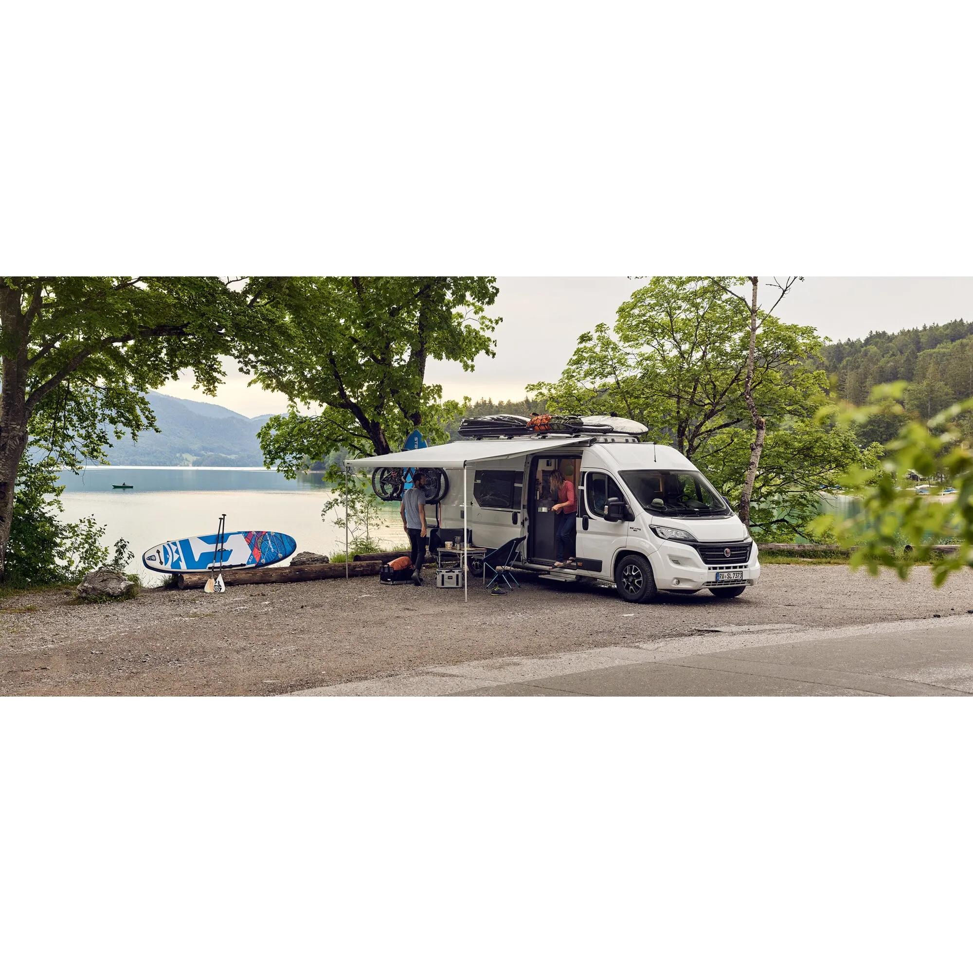 Weißer Fiat Ducato mit ausgefahrener Thule Omnistor 6300 Markise am See, daneben Fahrräder und ein Paddleboard