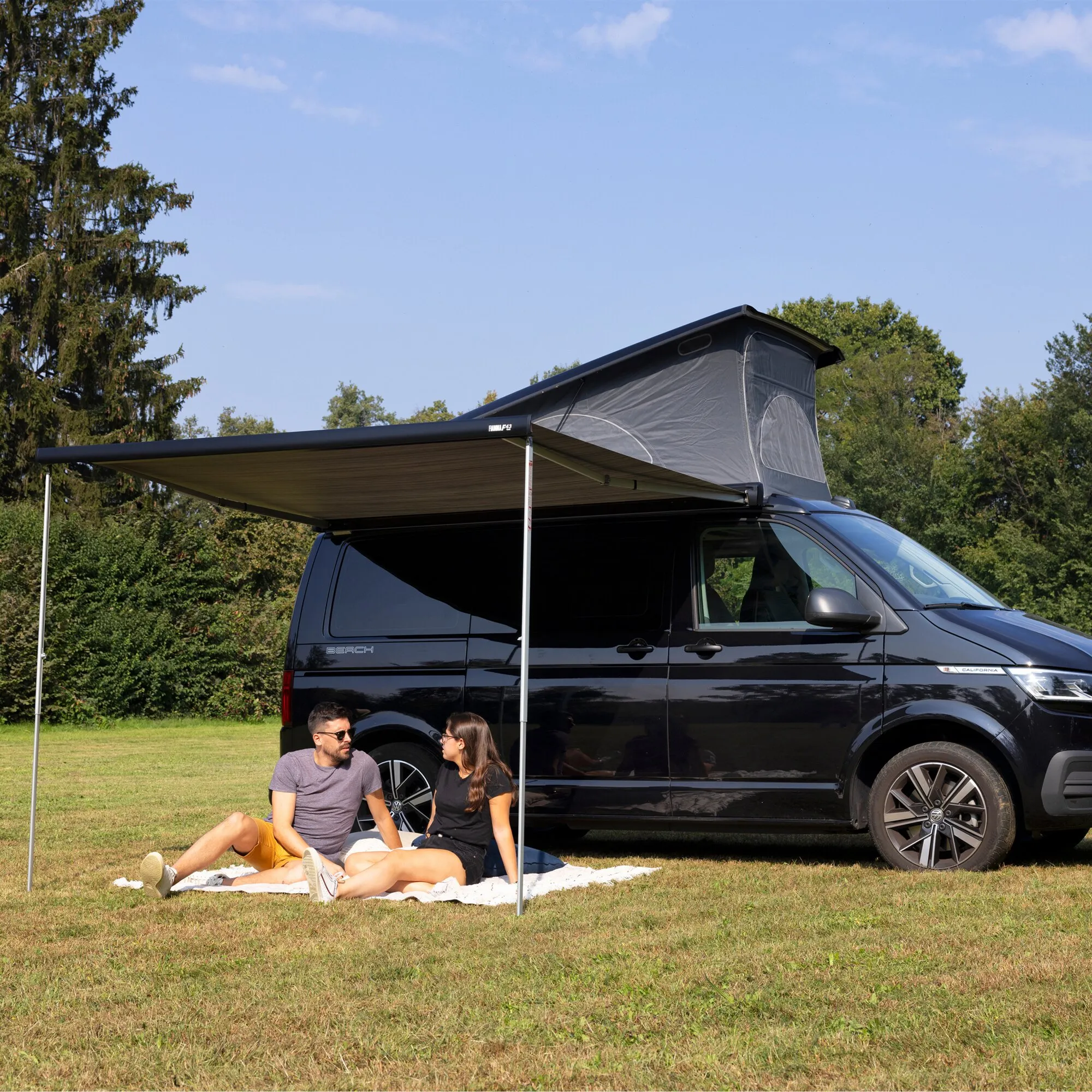 Schwarzer VW-Campingbus mit ausgefahrener Fiamma F43van-Markise und zwei sitzenden Personen auf einer Wiese