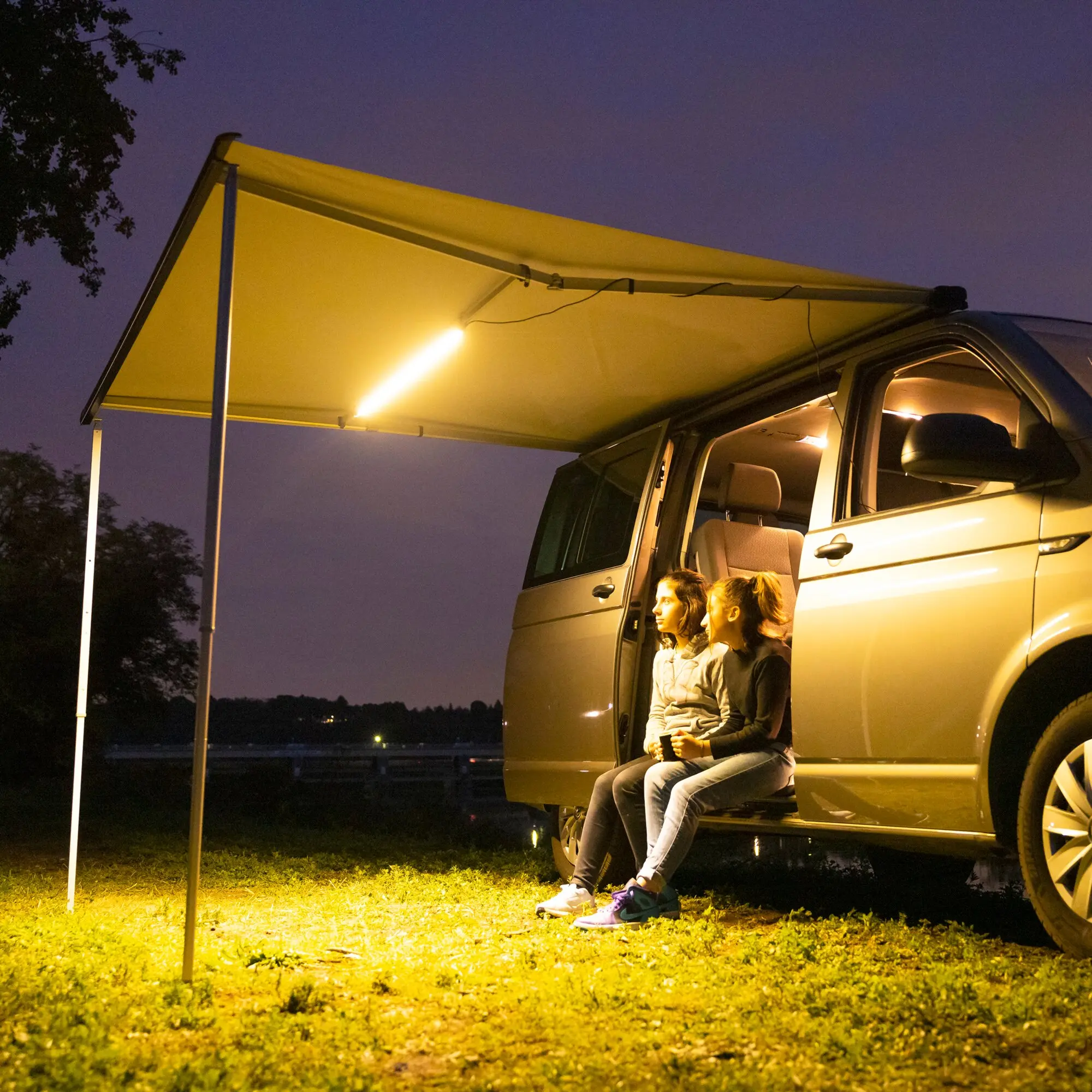 Nachtaufnahme eines weißen Vans mit ausgeklappter Seitenmarkise; LED-Lichtleiste leuchtet darunter, mehrere Jugendliche sitzen an der Tür