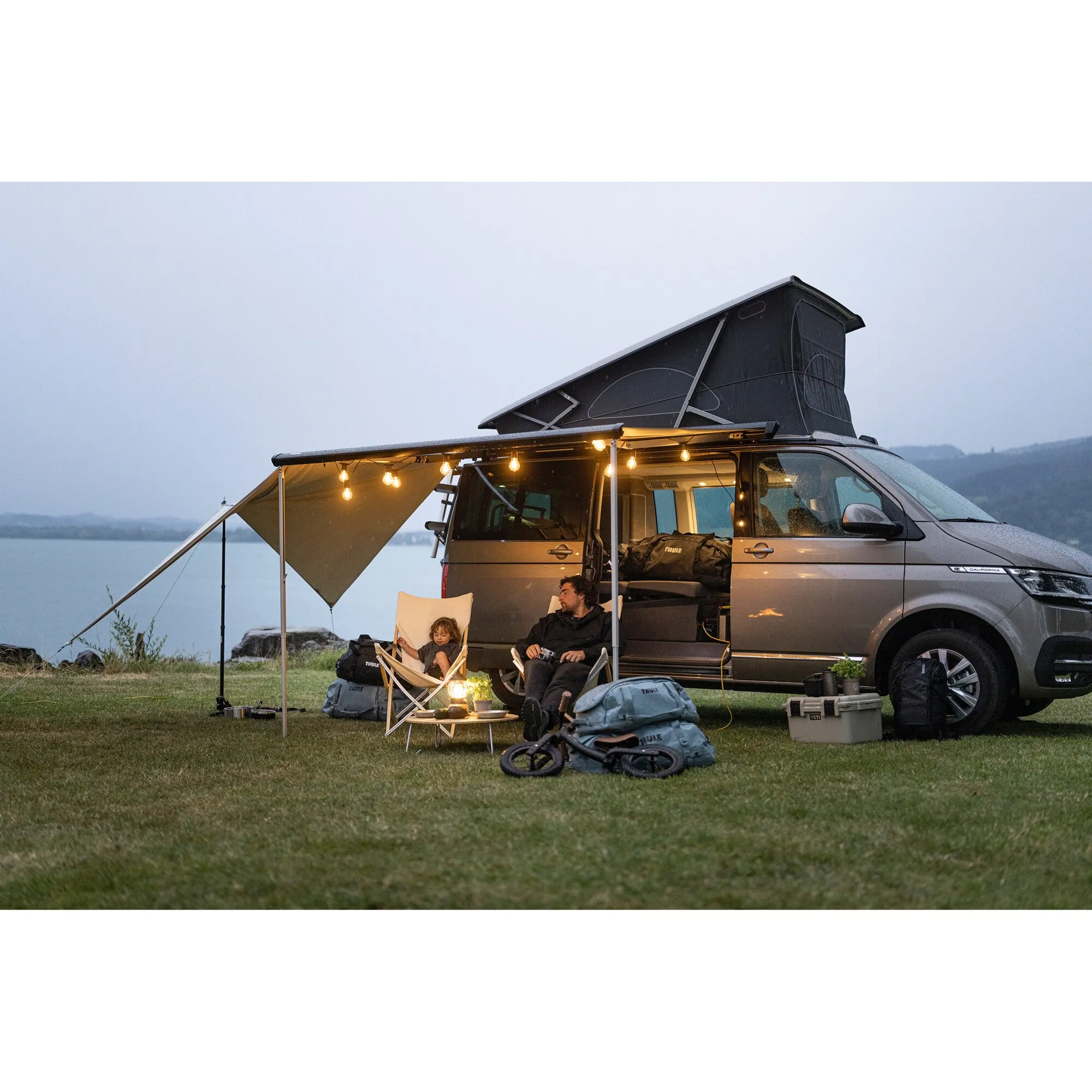 Grauer Van mit aufgeklapptem Dachzelt und Markise mit Lichterkette, davor Familie beim Campen, Gepäck und Fahrrad auf dem Gras am See