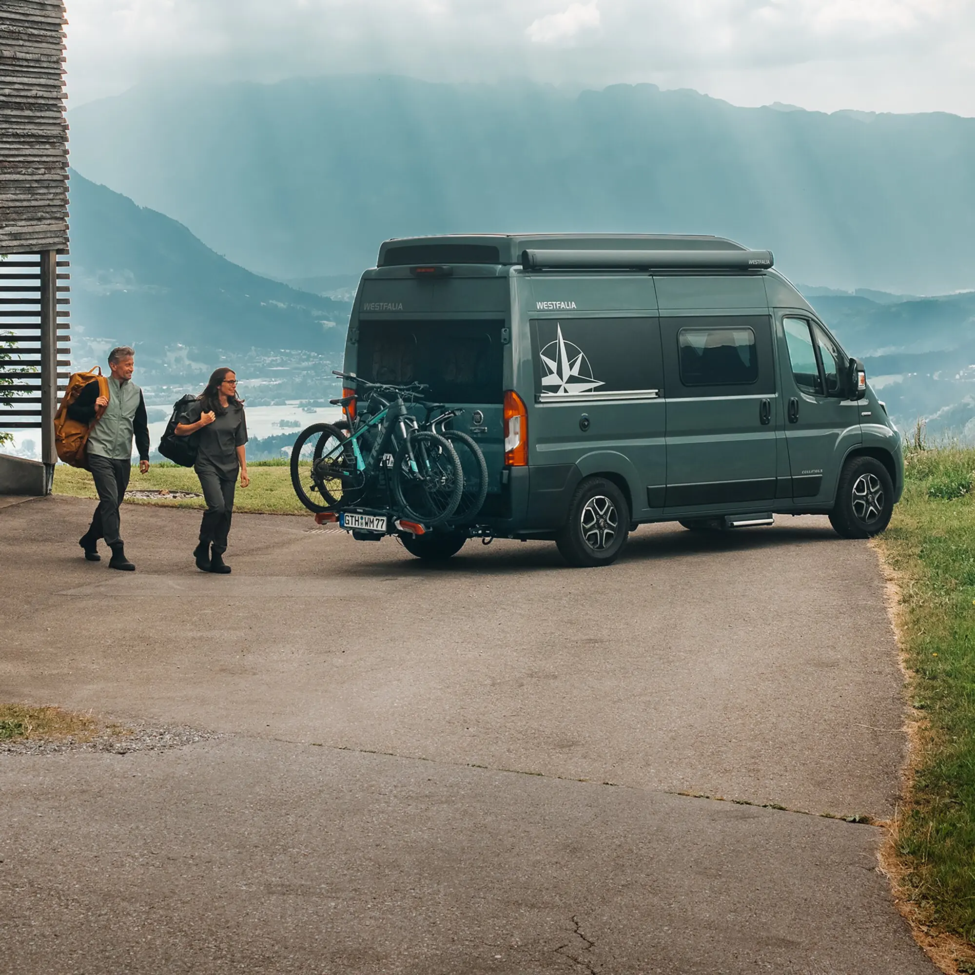 Zwei Fahrräder sind an einem Thule VeloSwing Heckträger an der Anhängerkupplung eines Vans montiert, davor zwei Personen mit Rucksäcken vor Berglandschaft