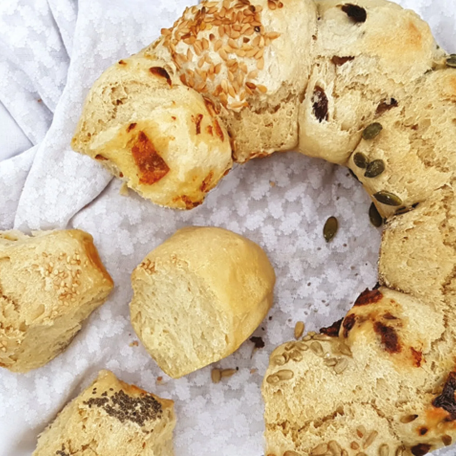 Verschiedene goldbraune Brotstücke mit Käse, Kräutern und Saaten auf einem weißen Küchentuch arrangementiert