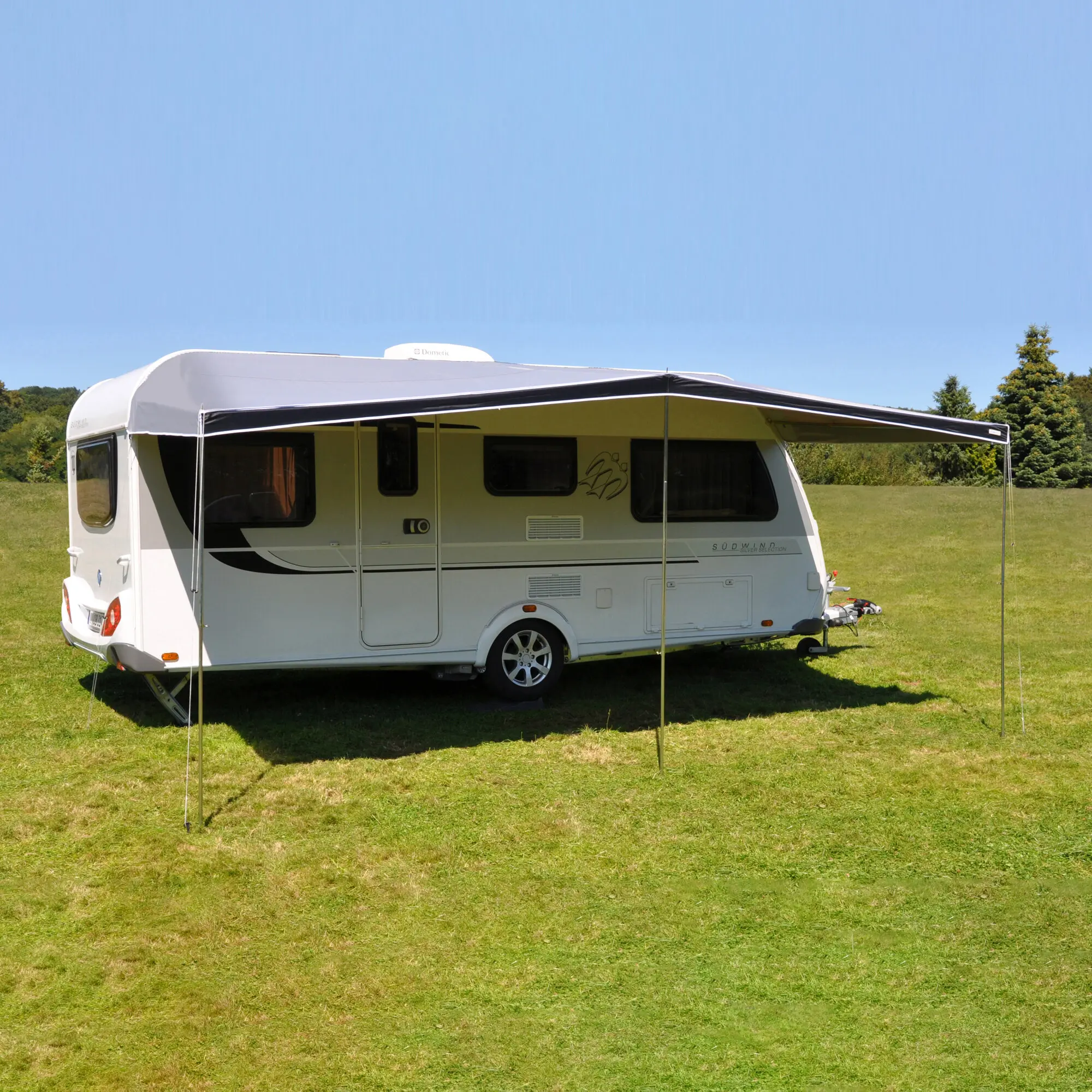 Weißes Reisemobil mit seitlich ausgefahrener Sonnendach-Kombi-Markise, Stützpfosten im Gras, grüne Wiese und blauer Himmel mit Bäumen im Hintergrund