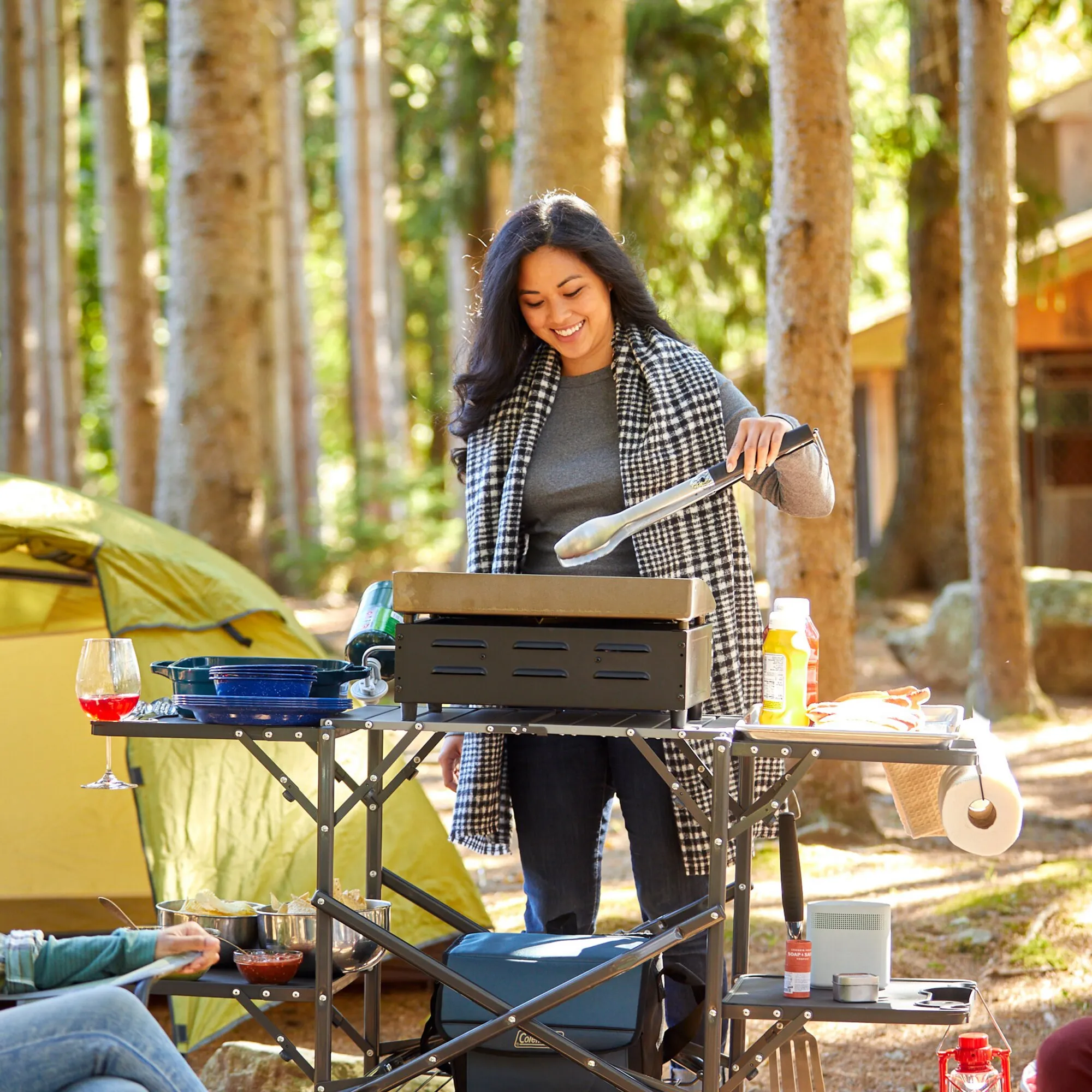 Frau bereitet am Grill der Campingküche Slim-Fold Cook Station™ im Wald Essen zu, mit Geschirr und Zutaten auf den Ablagen