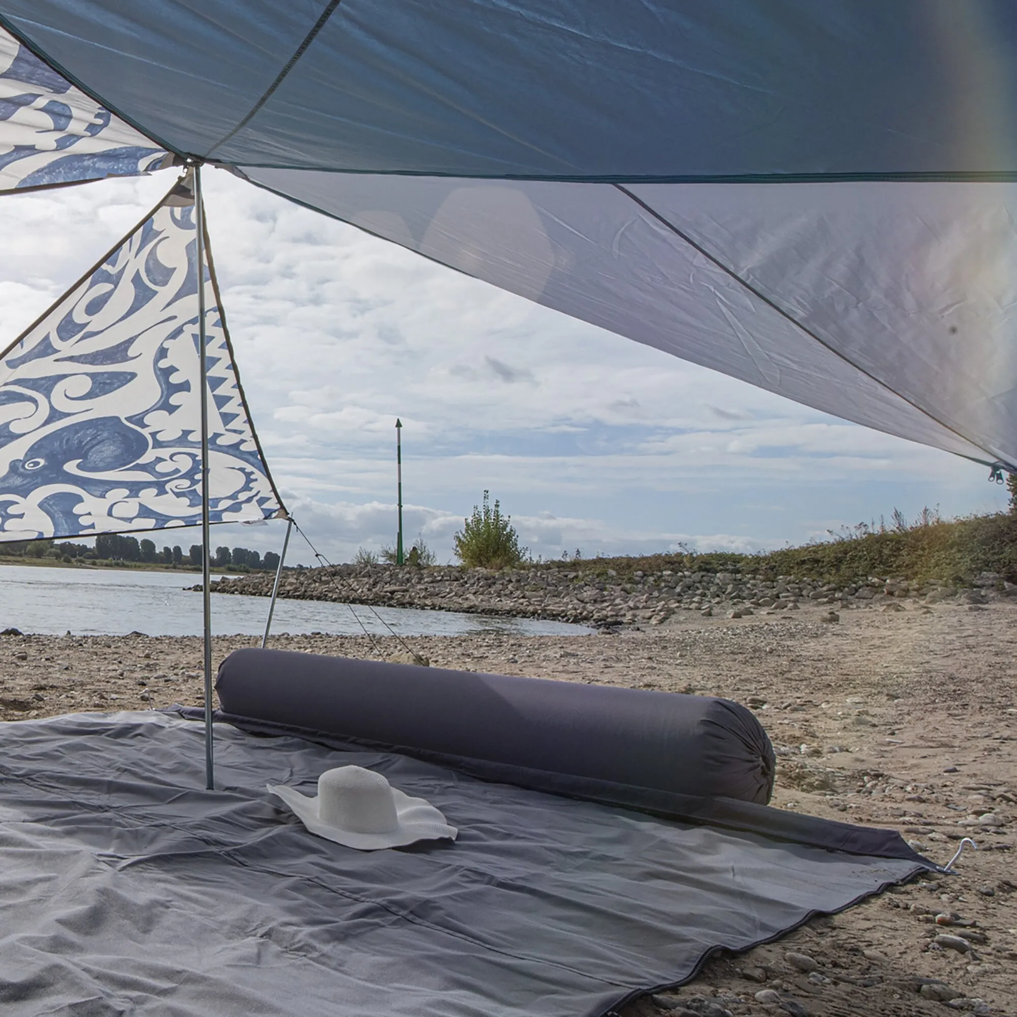 Dunkelgraue Kissenrolle „Zip XL Lounger“ liegt unter einem Sonnensegel am steinigen Ufer neben einer Picknickdecke