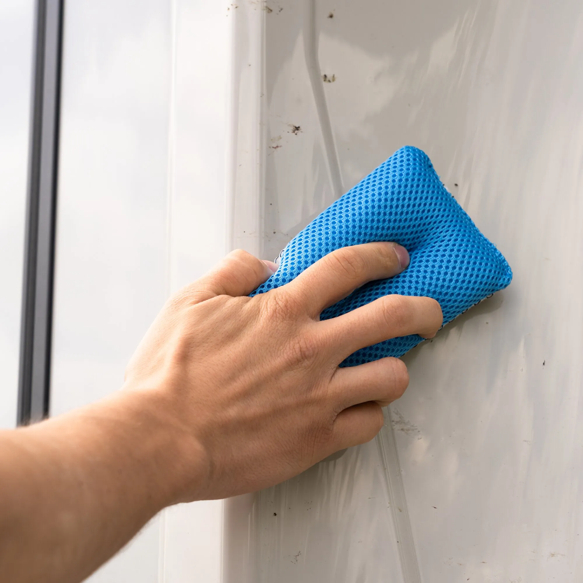 Hand hält einen blauen Bug Buster Schwamm mit Wabenstruktur und reinigt damit verschmutzten Fensterrahmen mit Insektenresten