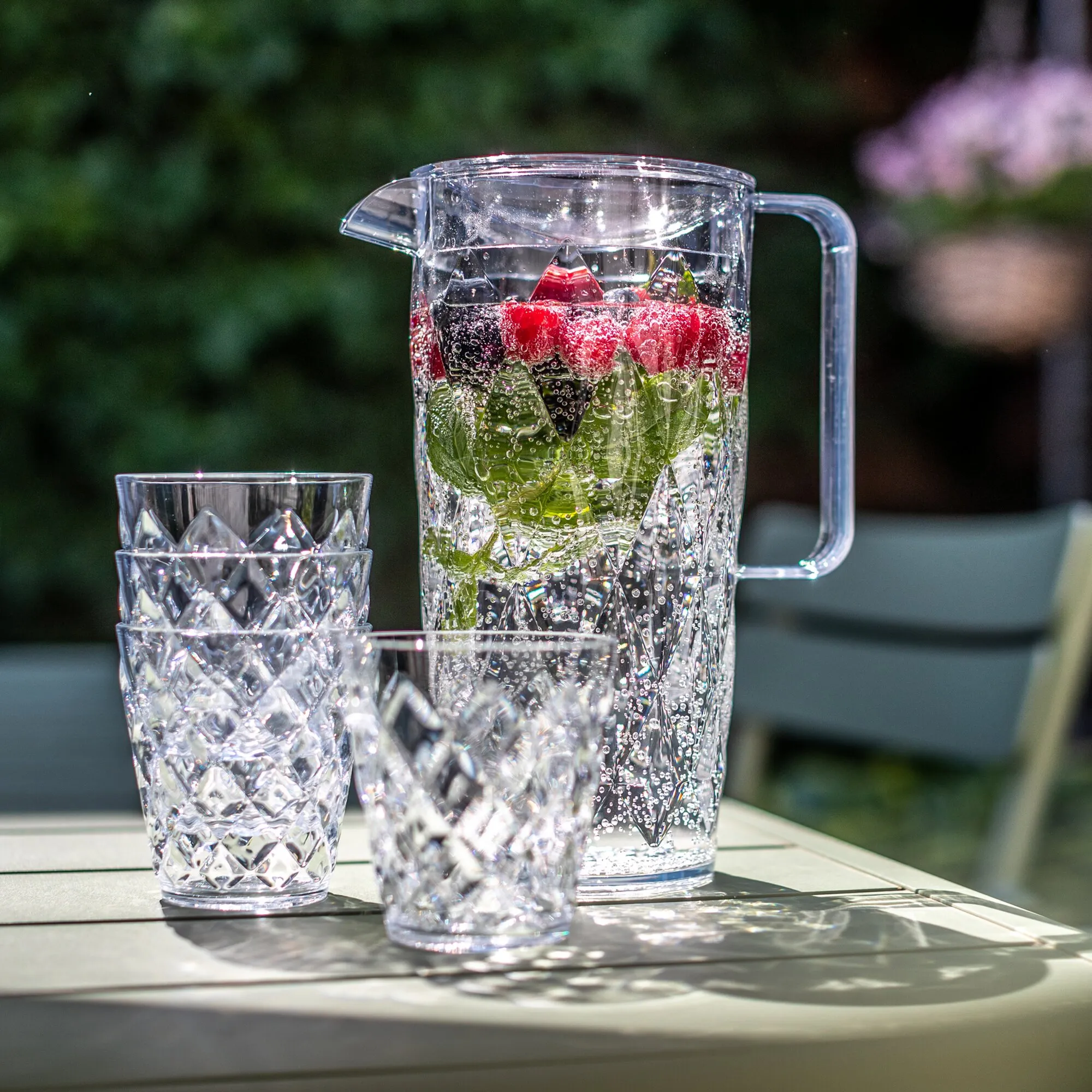 Transparente CRYSTAL-Kanne mit vier strukturierten Bechern, gefüllt mit Wasser, Erdbeeren und Minze auf einem Gartentisch