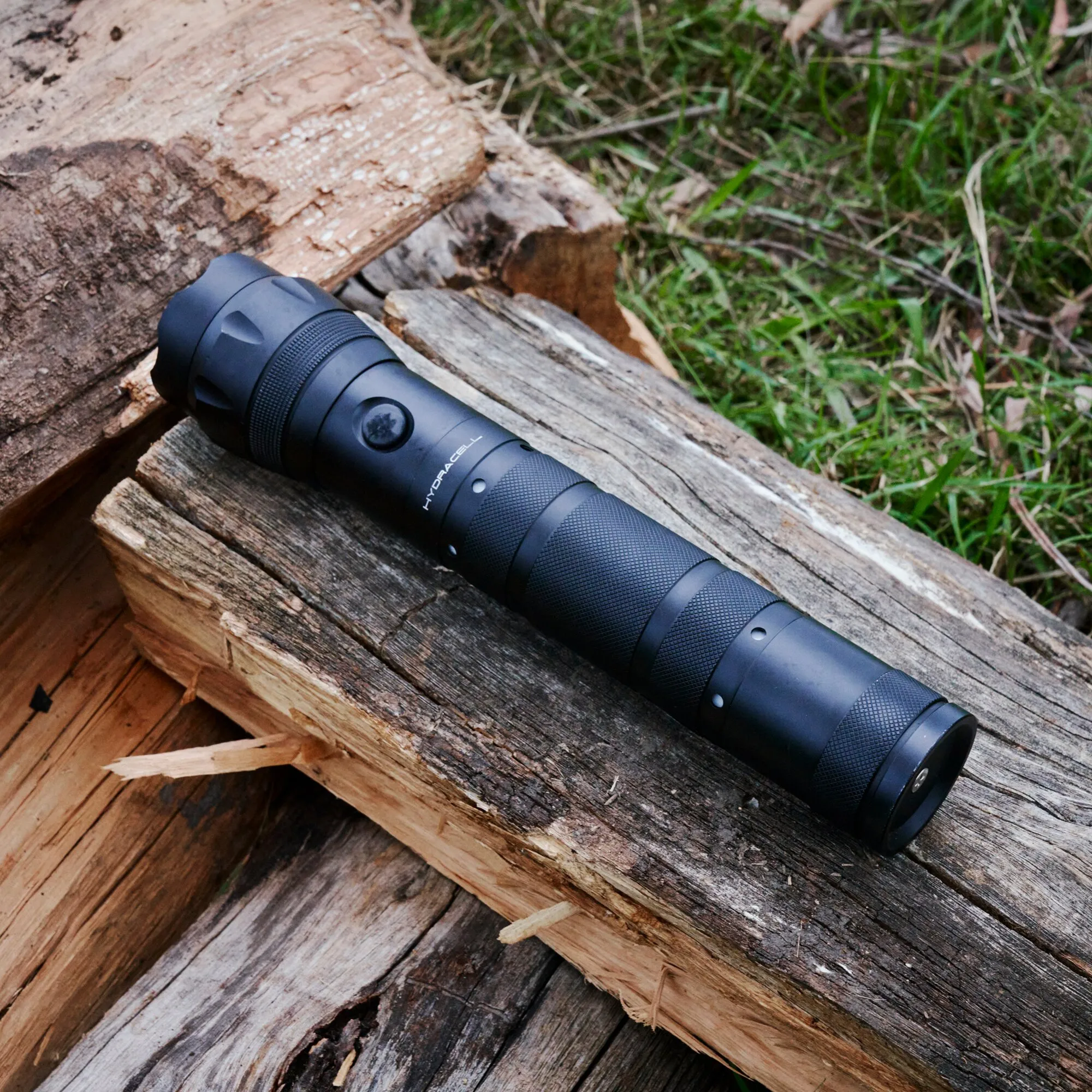 Schwarze Taschenlampe AQUA PRO von Hydracell mit geriffeltem Griff liegt auf verwitterten Holzstämmen im Freien