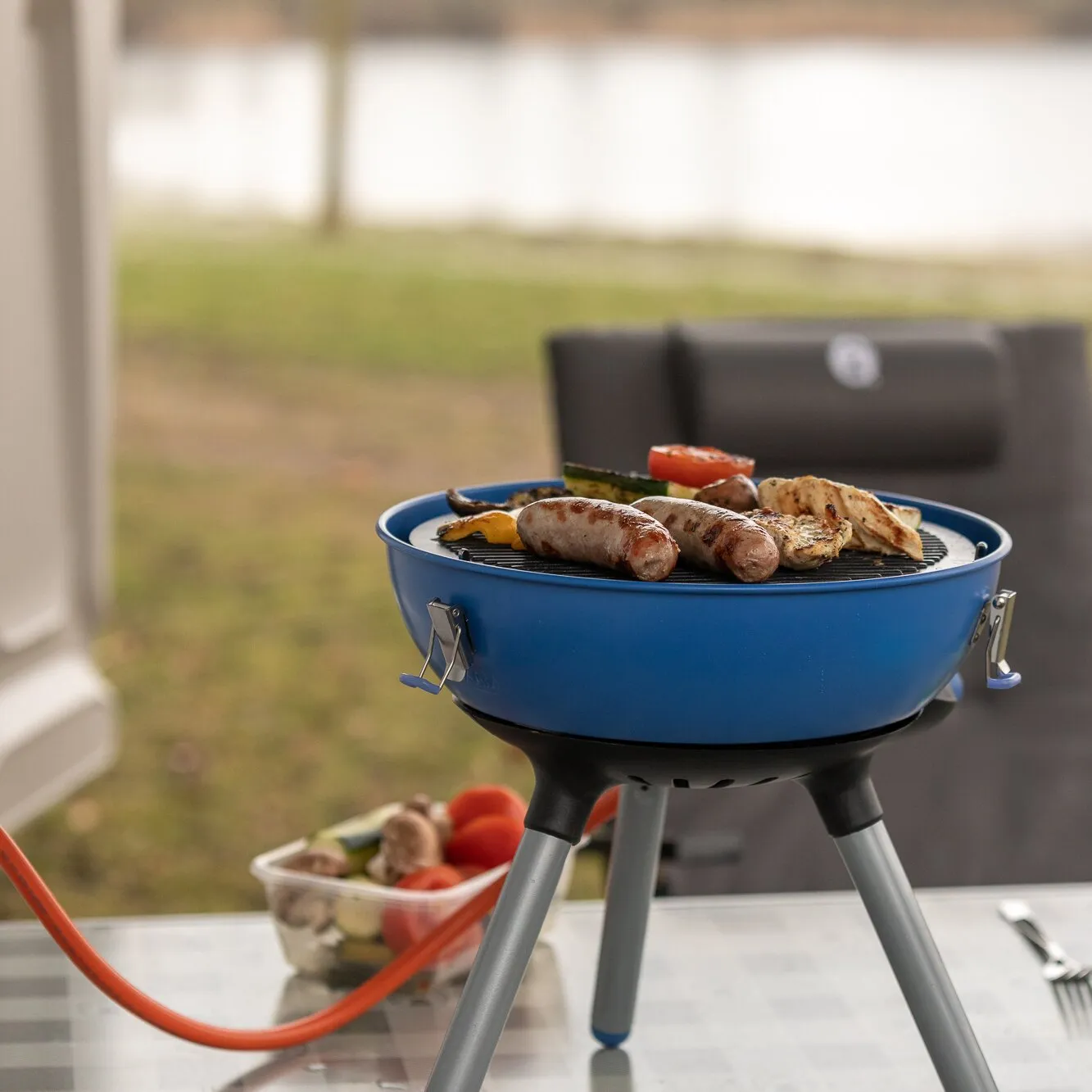Blauer Party Grill 400 Caravan Connect auf Standbeinen mit Würstchen, Gemüse und Hähnchen auf dem Grillrost vor einem Wohnmobil