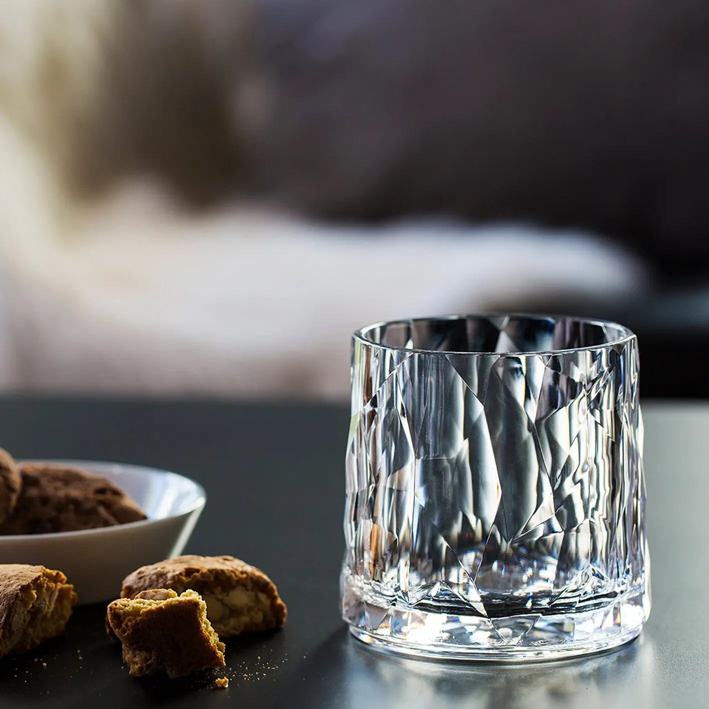 Nahaufnahme eines klaren, facettierten Whiskyglases aus Kristall mit prismatischen Mustern, daneben liegen Biscotti auf einer dunklen Tischplatte