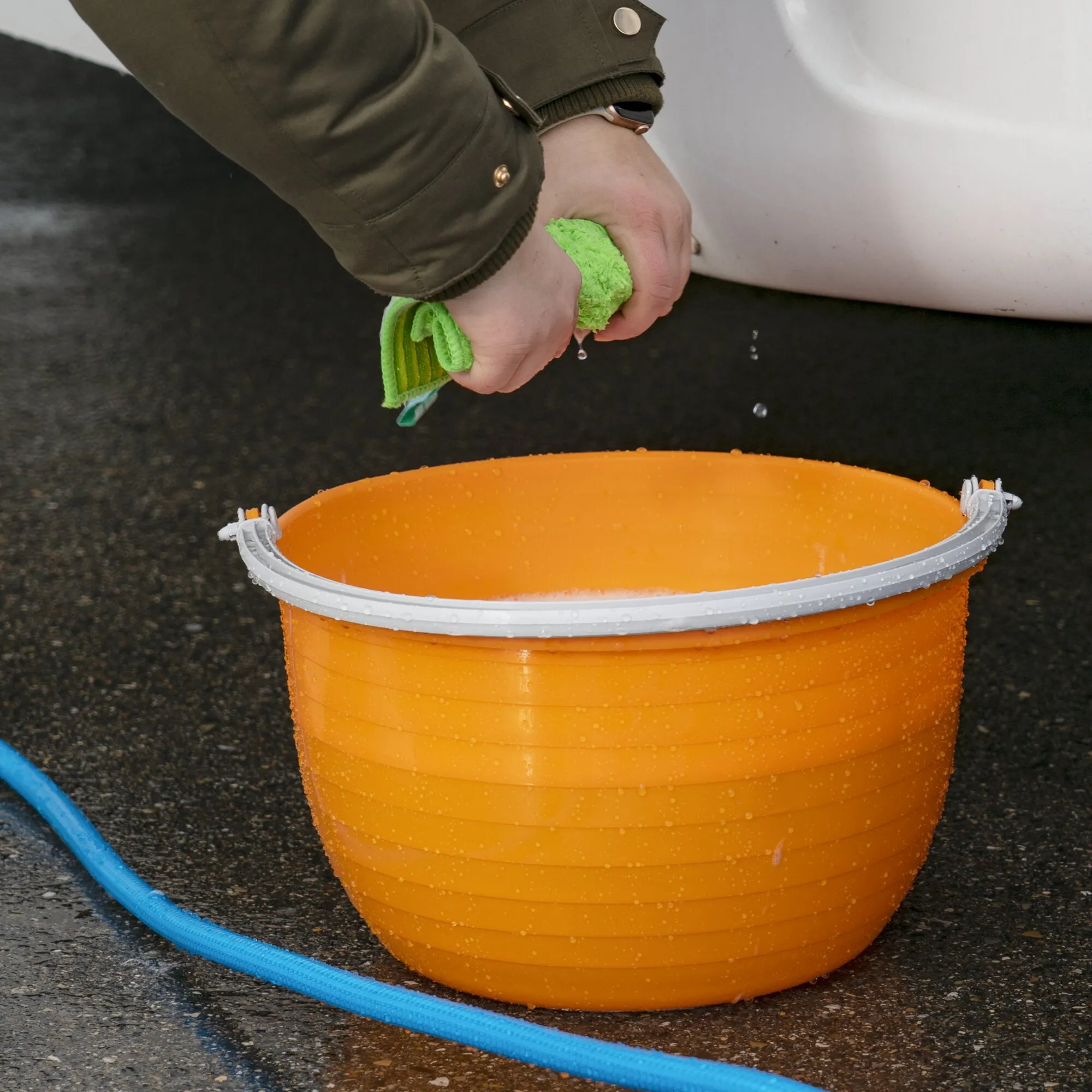 Orangefarbener Vielzweckeimer Vinis mit weißem Tragebügel beim Auswringen eines grünen Reinigungstuchs, von Wassertropfen umgeben