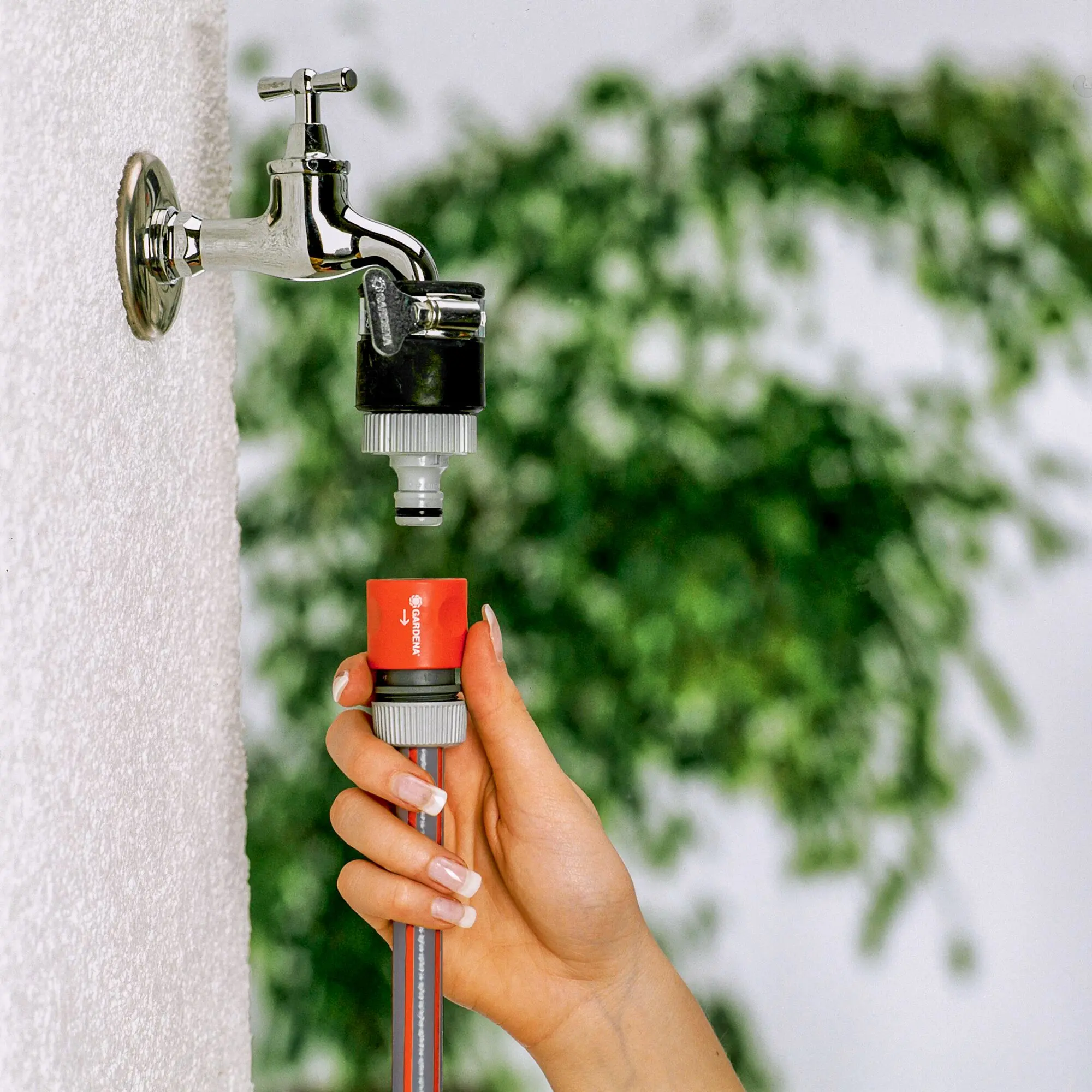 Nahaufnahme eines glänzenden Außenwasserhahns, an dem eine Hand mit lackierten Nägeln einen roten Gardena-Schnellverbinder am Schlauch befestigt