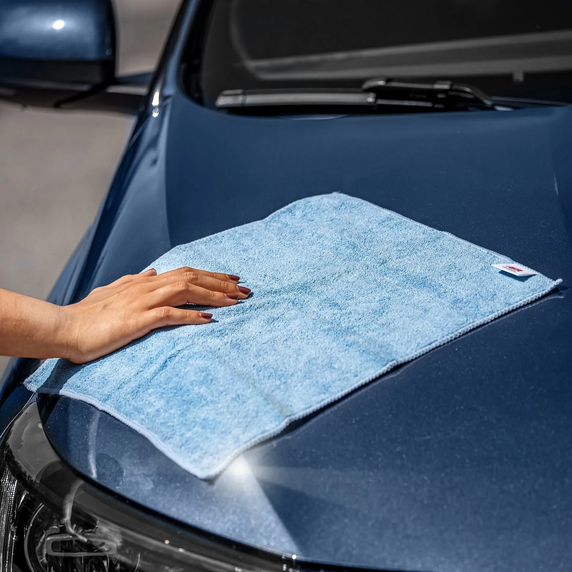 Hand wischt mit einem hellblauen Mikrofaser-Reinigungstuch über die Motorhaube eines dunkelblauen Autos