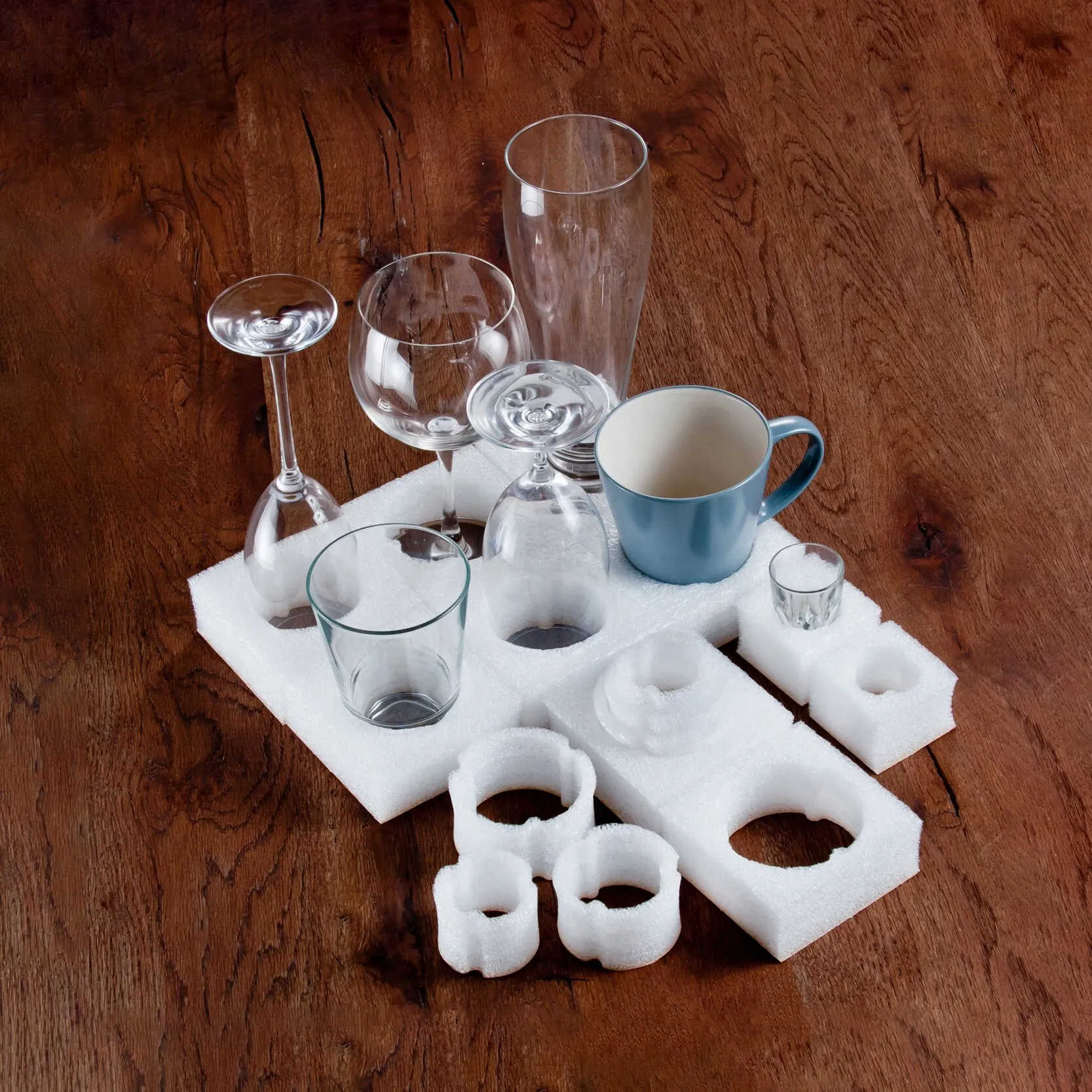 Weißes Glassicherungstray aus Schaumstoff mit Aussparungen für verschiedene Gläser und eine blaue Tasse auf Holztisch