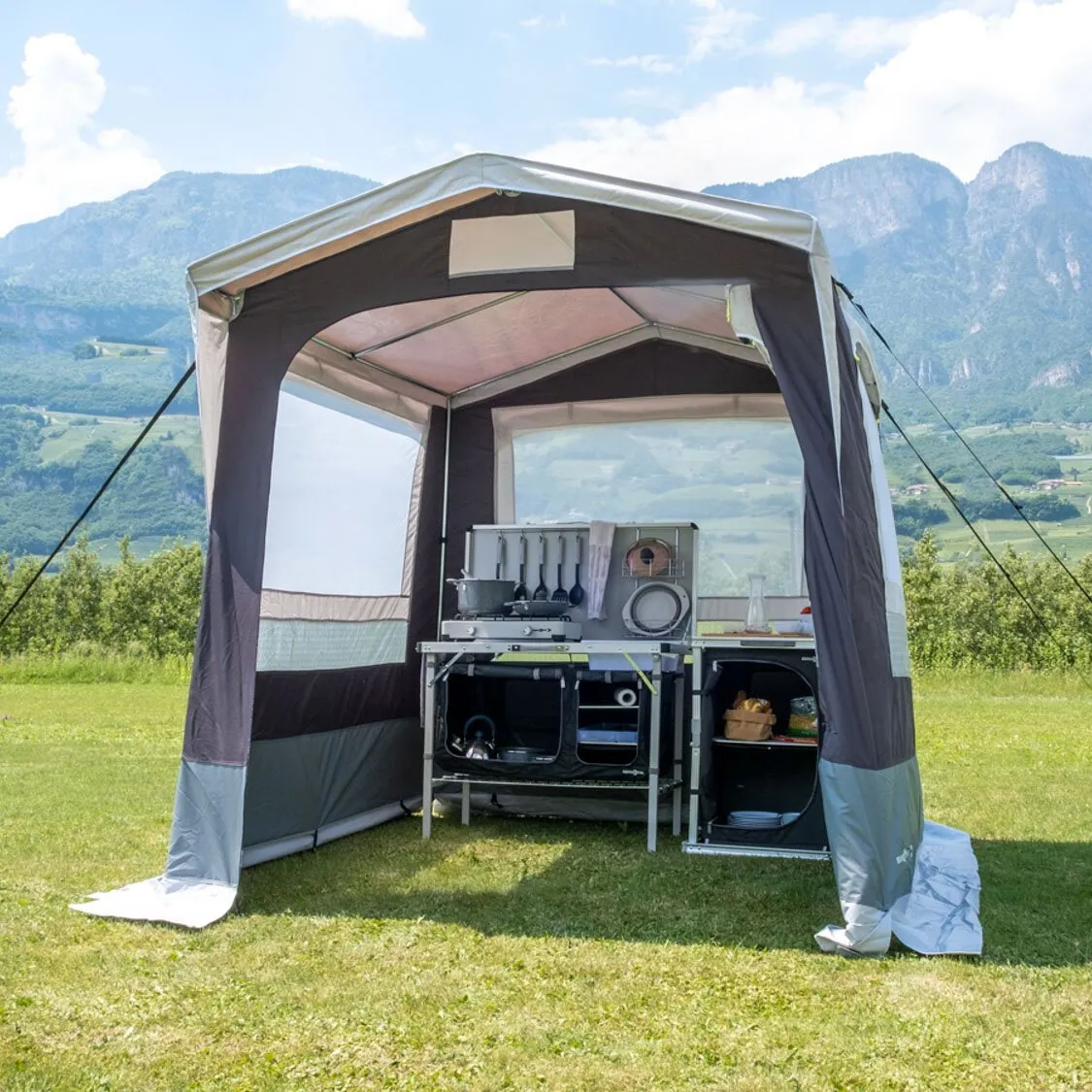 Dunkelgraues Küchenzelt Gusto II NG mit aufgebauter Campingküche auf einer Wiese vor Berglandschaft