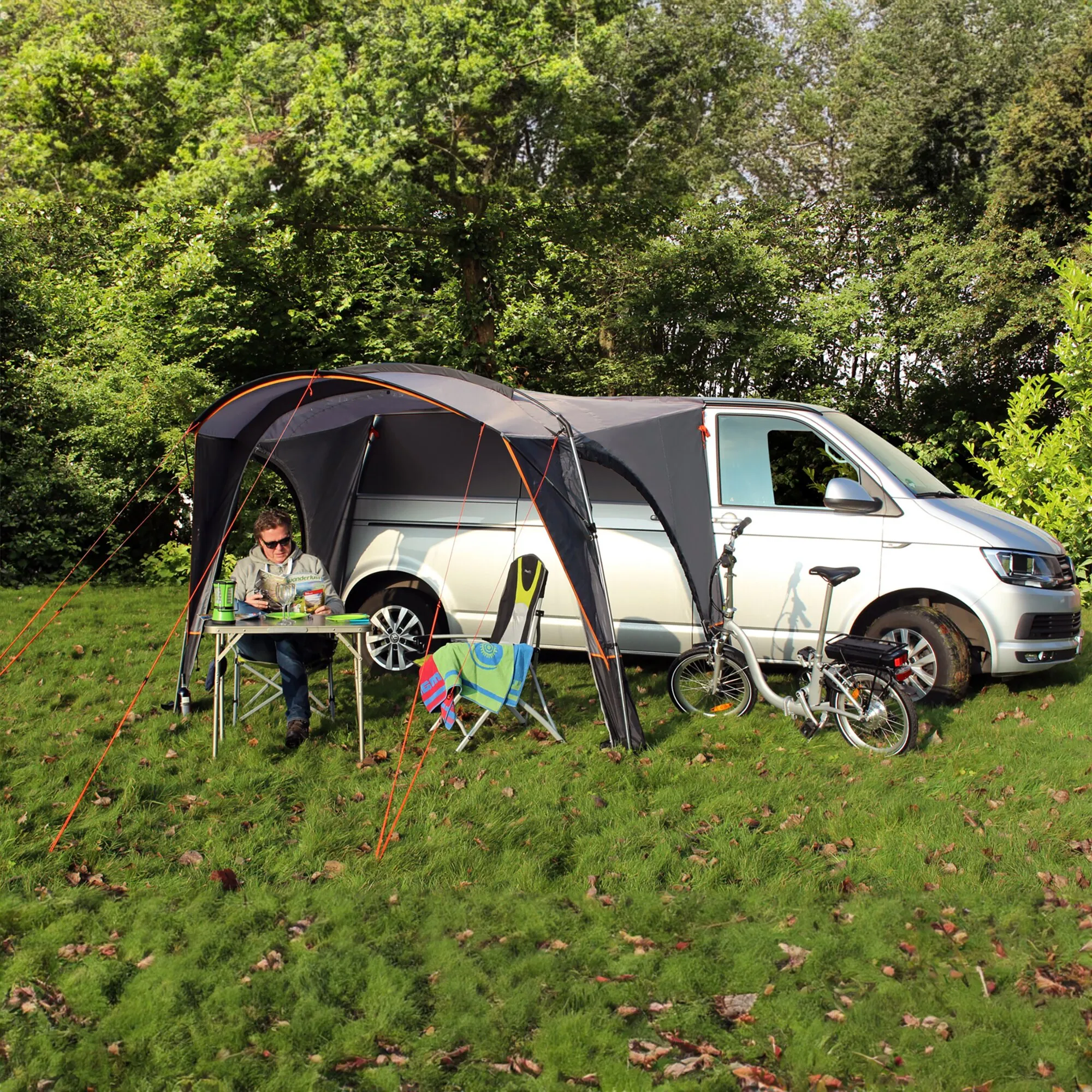 Busvorzelt Pisa in Schwarz und Grau, an einem weißen Campervan befestigt, mit Tisch, Campingstuhl und Fahrrädern auf einer Wiese、】【