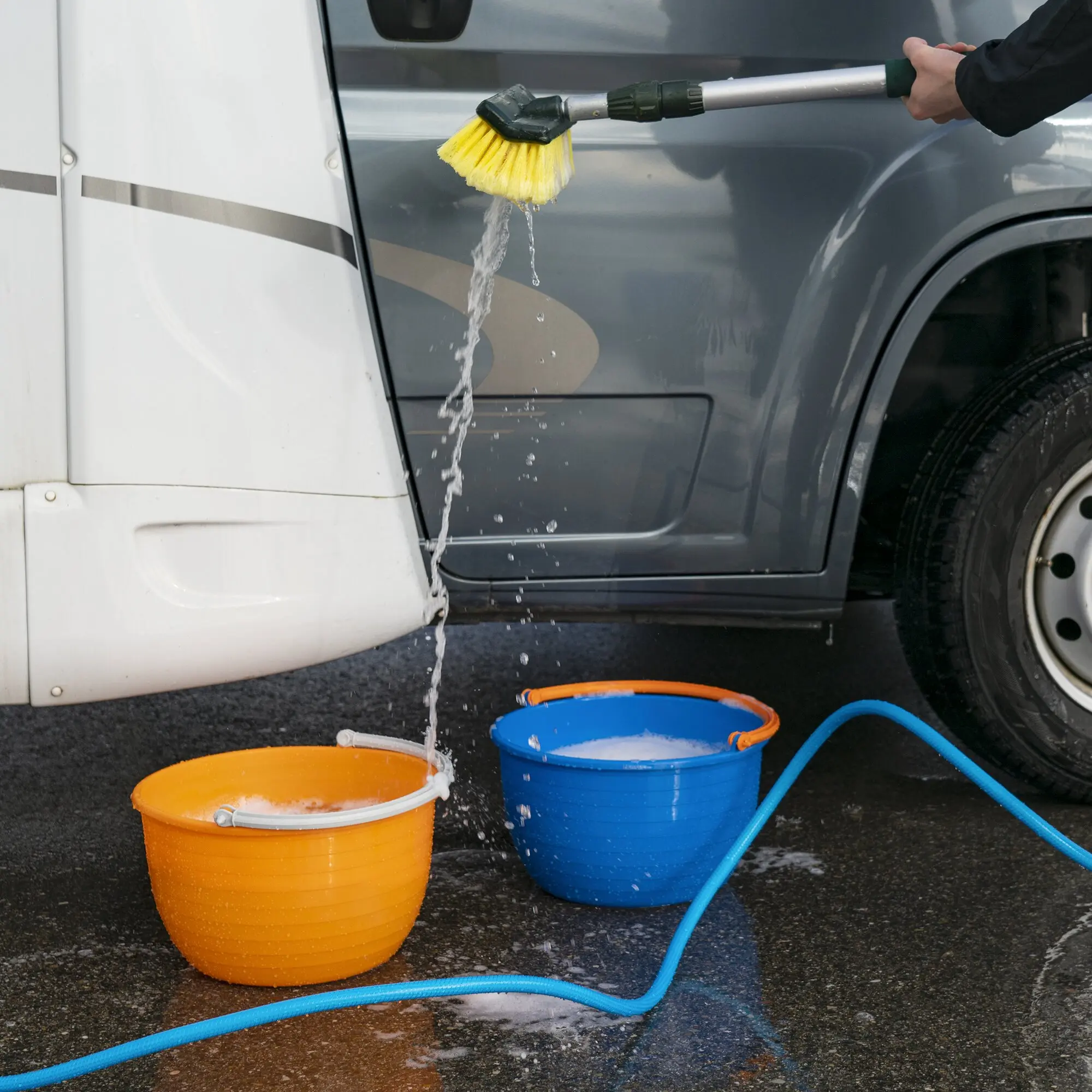 Orangefarbener Vielzweckeimer Vinis steht neben einem blauen Eimer auf nassem Asphalt, während eine gelbe Bürste mit blauem Schlauch zum Autowaschen verwendet wird