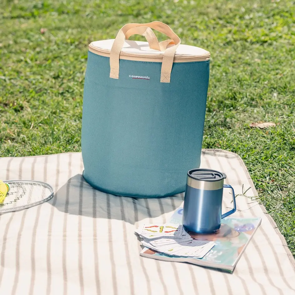 Kühltasche Fold‘N Cool 15L in blauem Teal mit beigefarbenen Griffen steht auf einer Picknickdecke im Gras, daneben blaue Thermoskanne, Karten und eine Zeitschrift, Rand eines Badmintonschlägers sichtbar