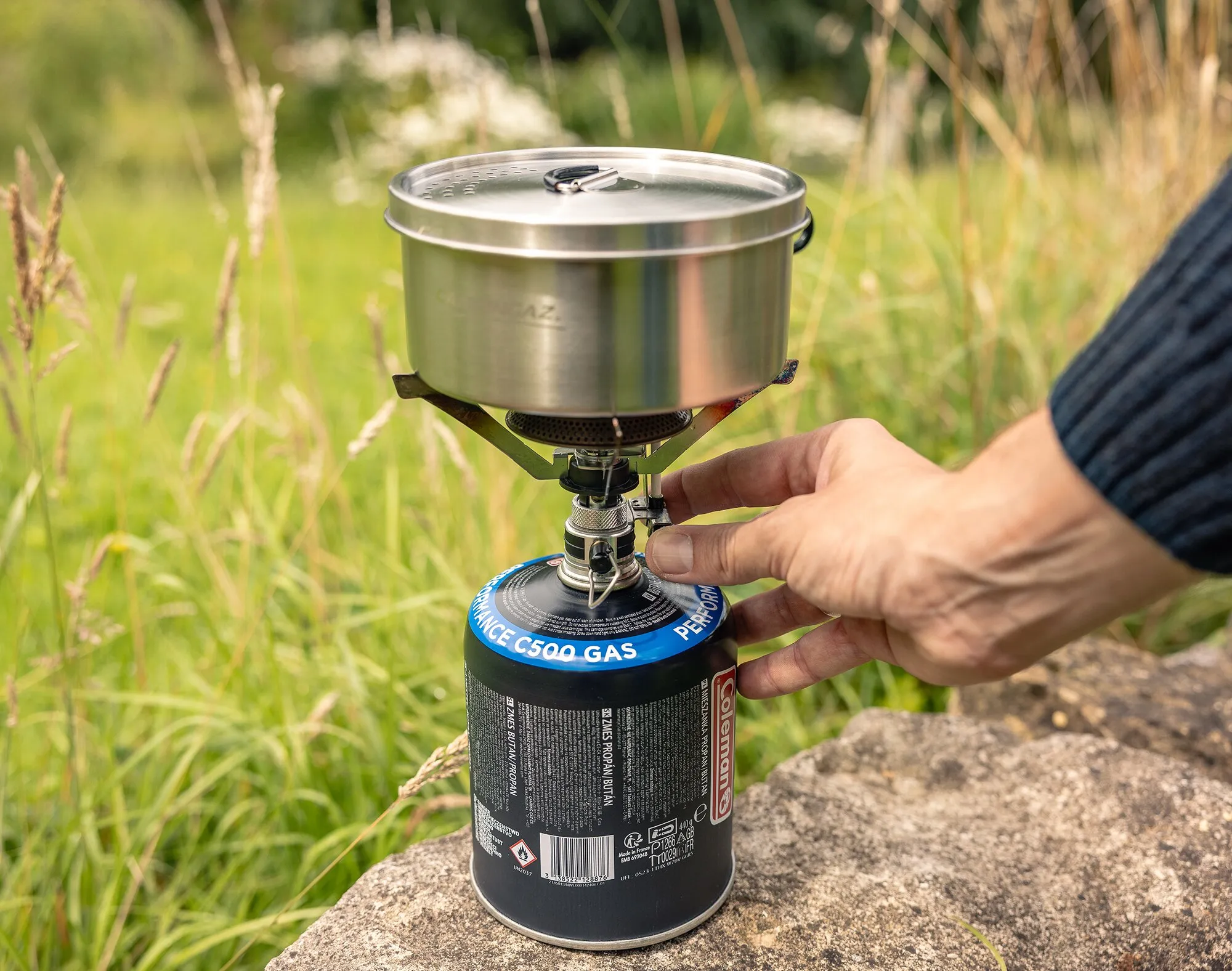Edelstahl-Trekking-Kochgeschirr mit Deckel auf einem Campingkocher, während eine Hand die Gaskartusche im Freien bedient