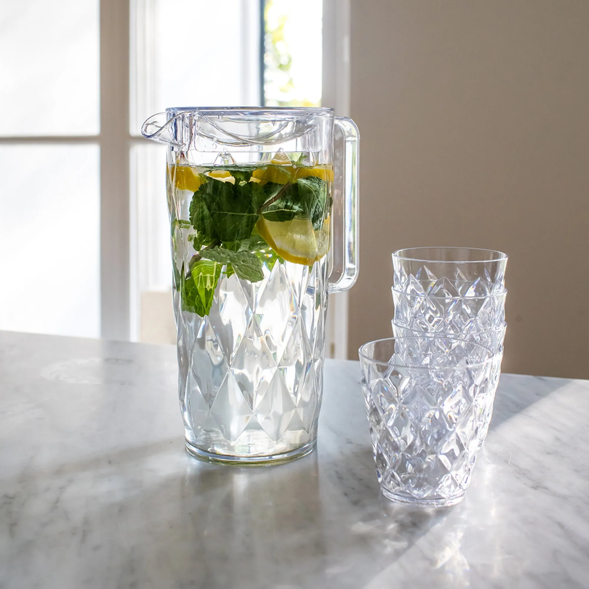 Transparente Kanne CRYSTAL mit strukturiertem Muster, Zitronen- und Minzwasser sowie vier passende Becher auf Marmortisch