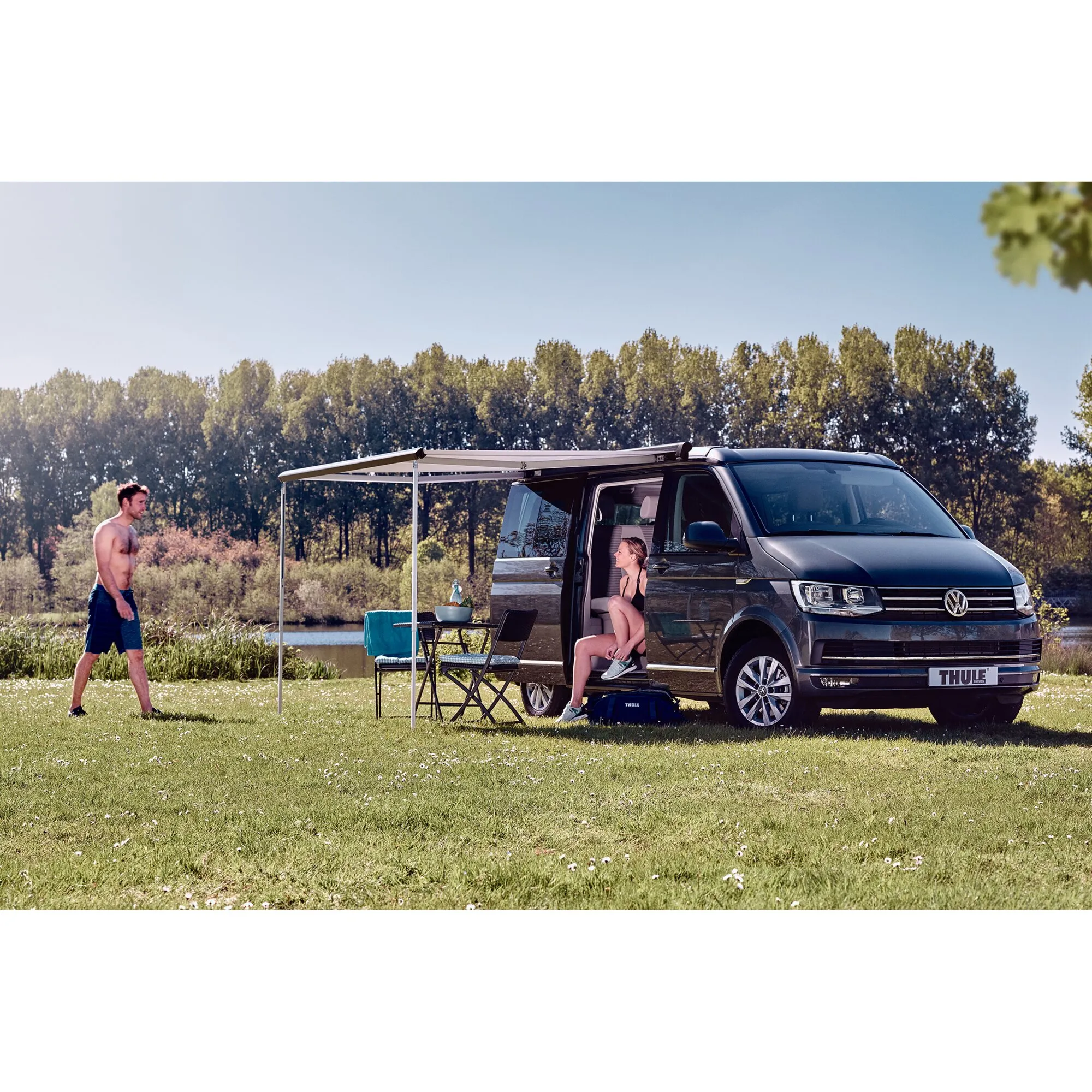 Anthrazitfarbene Thule Omnistor 5102 Markise an einem Volkswagen-Bus auf einer Wiese am See mit Sitzgruppe und zwei Personen