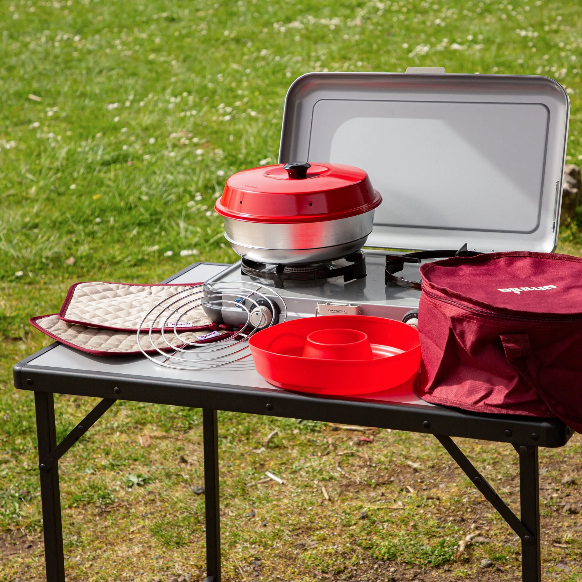 Roter Campingbackofen mit Deckel auf einem Gaskocher, dazu Backblech, Grillrost, Backform und Transporttasche auf einem Campingtisch im Freien