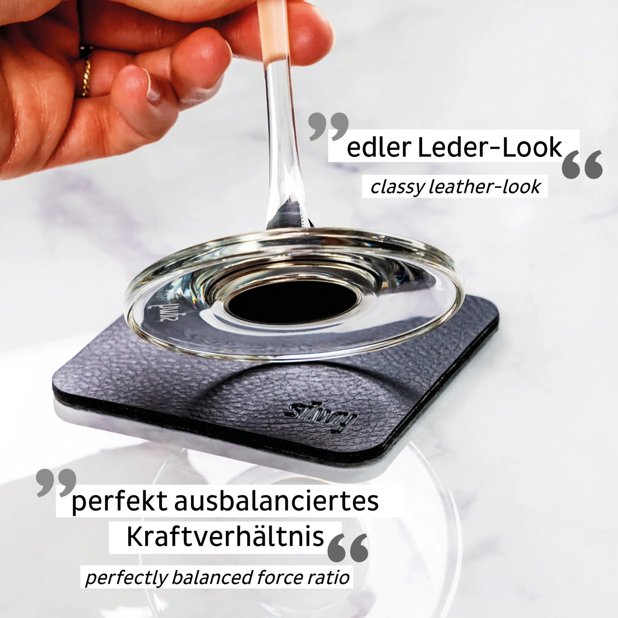 Hand hält ein transparentes Kristall-Champagnerglas an einem schlanken Stiel über einem runden schwarzen Magnet-Untersetzer in Lederoptik