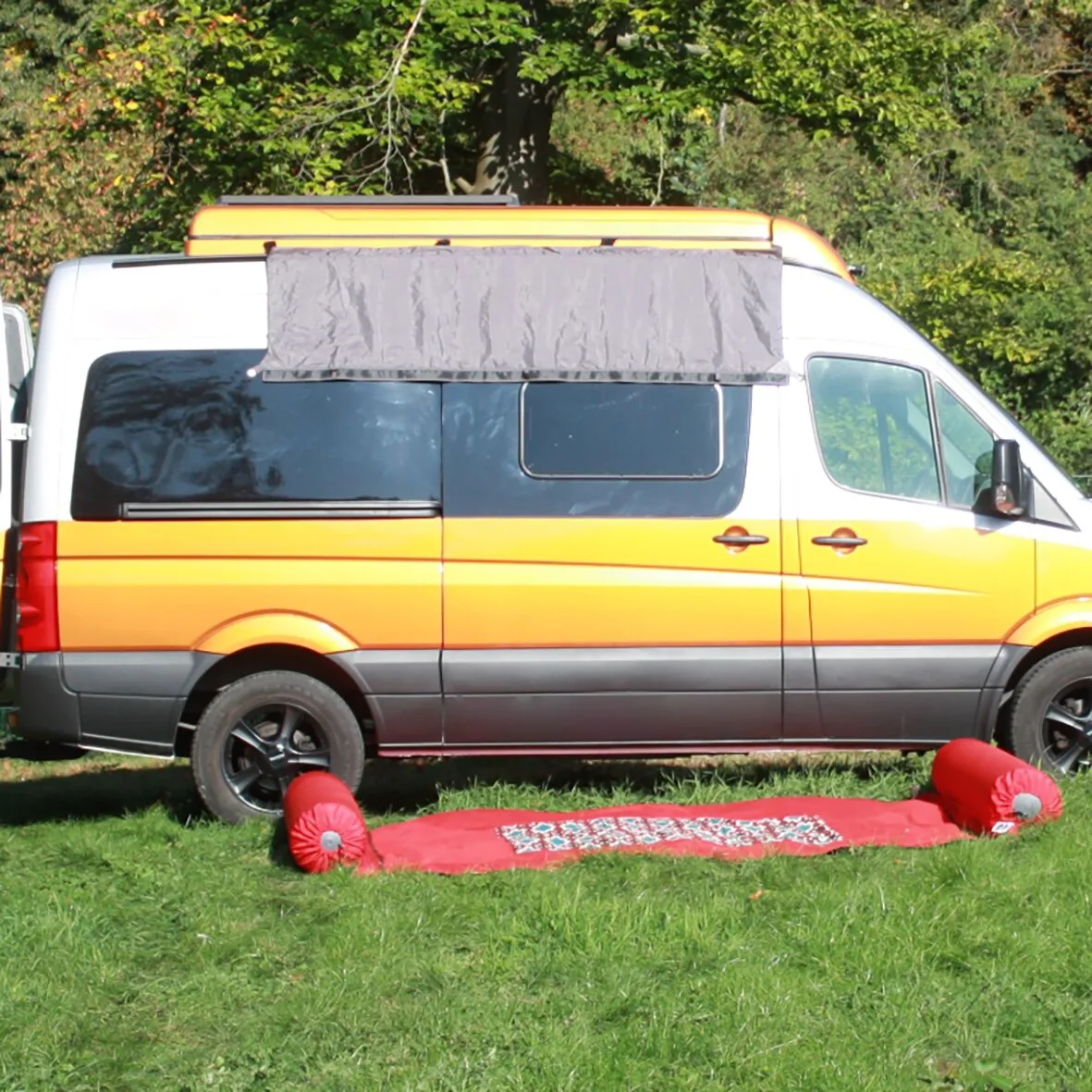 Universal Fahrzeugadapter XL als graue Stoffverbindung am Dach eines orange-weißen Campingvans in einer Waldumgebung