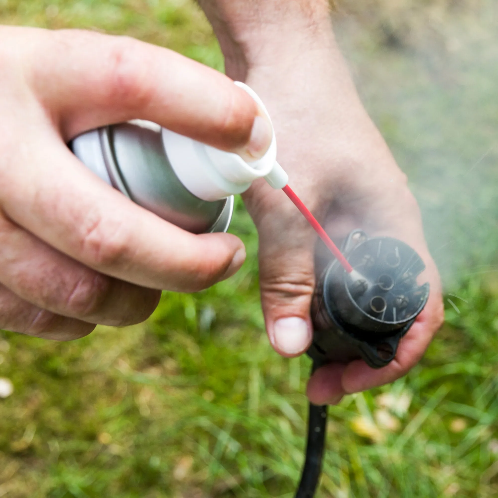 Hand sprüht Multifunktionsspray GT7 mit rotem Sprühröhrchen auf ein schwarzes Gartengerät im Grünen