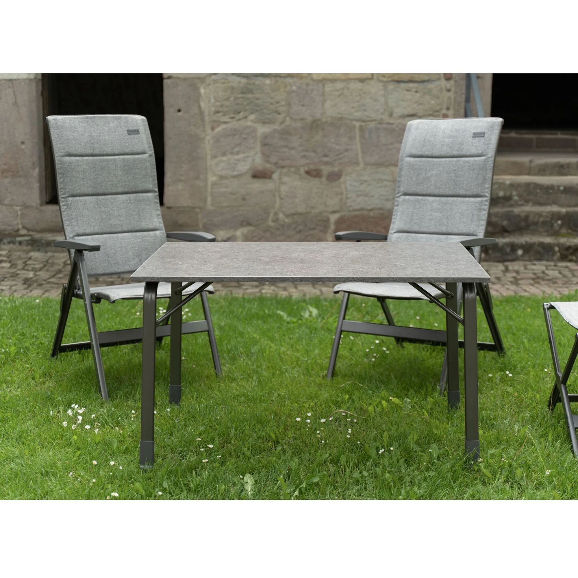 Zwei graue Campingstühle Elegance SG mit Armlehnen stehen auf einer Wiese an einem Gartentisch vor einer Steinmauer