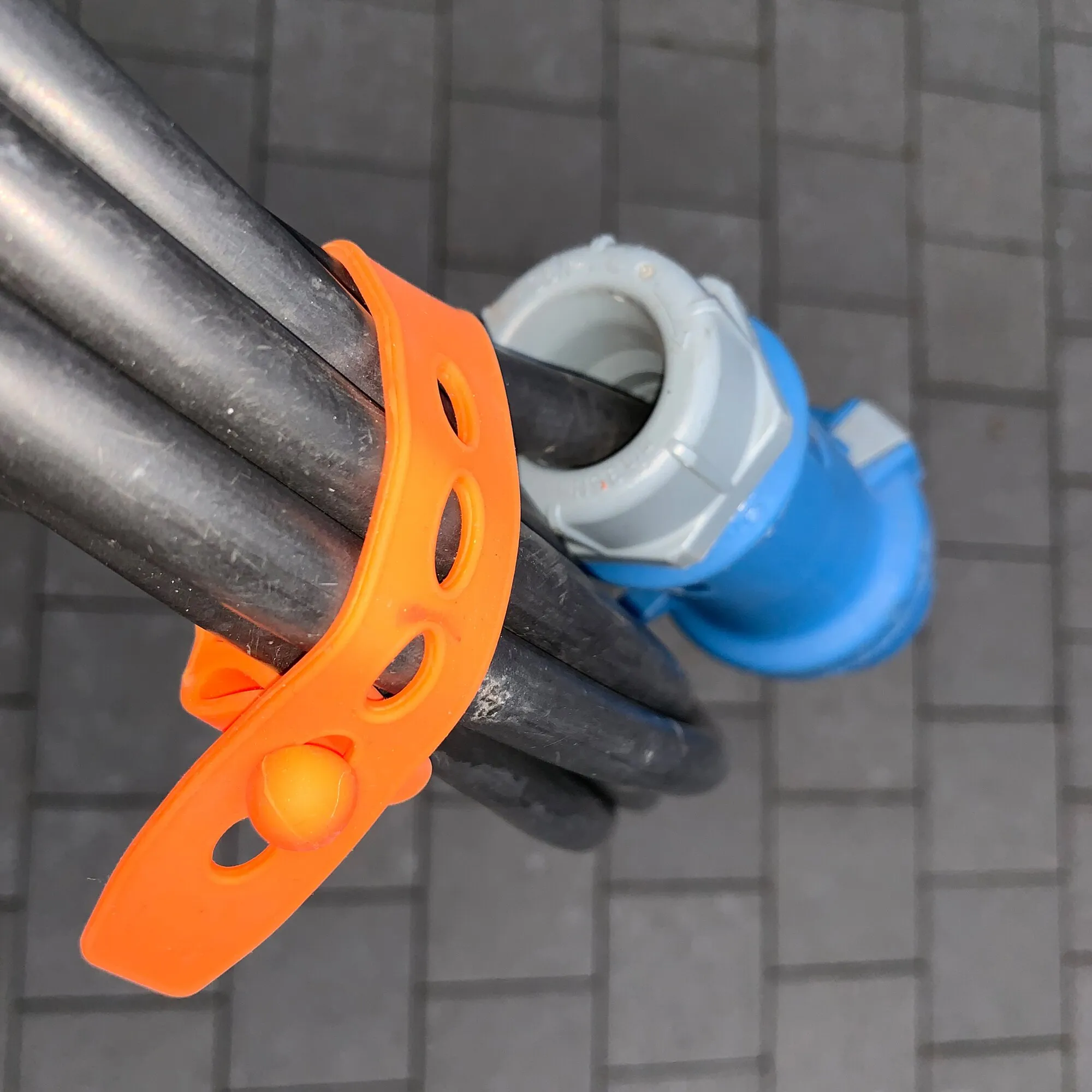 Orangefarbener PP-Silikonriemen mit mehreren Durchstecköffnungen, um schwarze Rohre gelegt; dahinter blaue und weiße Riemen an einem Gitter 
