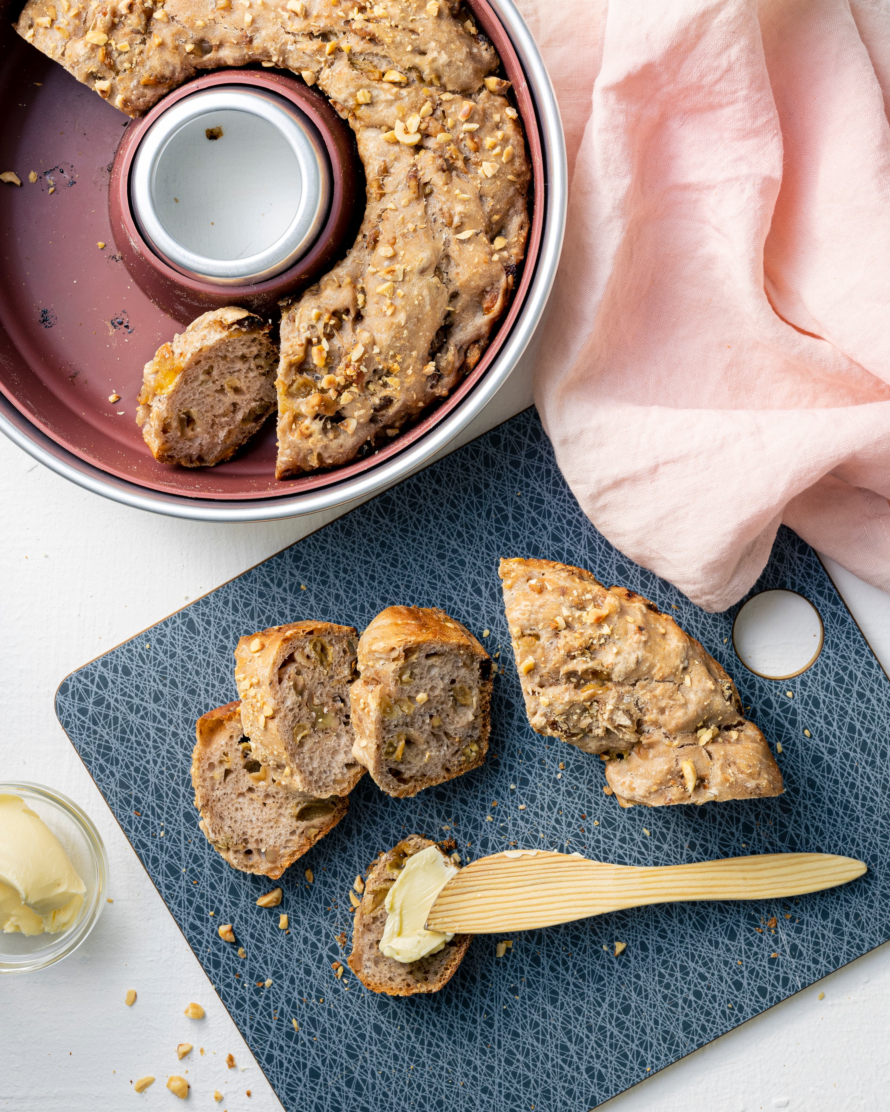 Zwei Silikonformen, rund, Rosé, mit Metallring, darin gebackenes Brot mit Nüssen, Scheiben daneben, Butter, Holzspatel und pinke Serviette