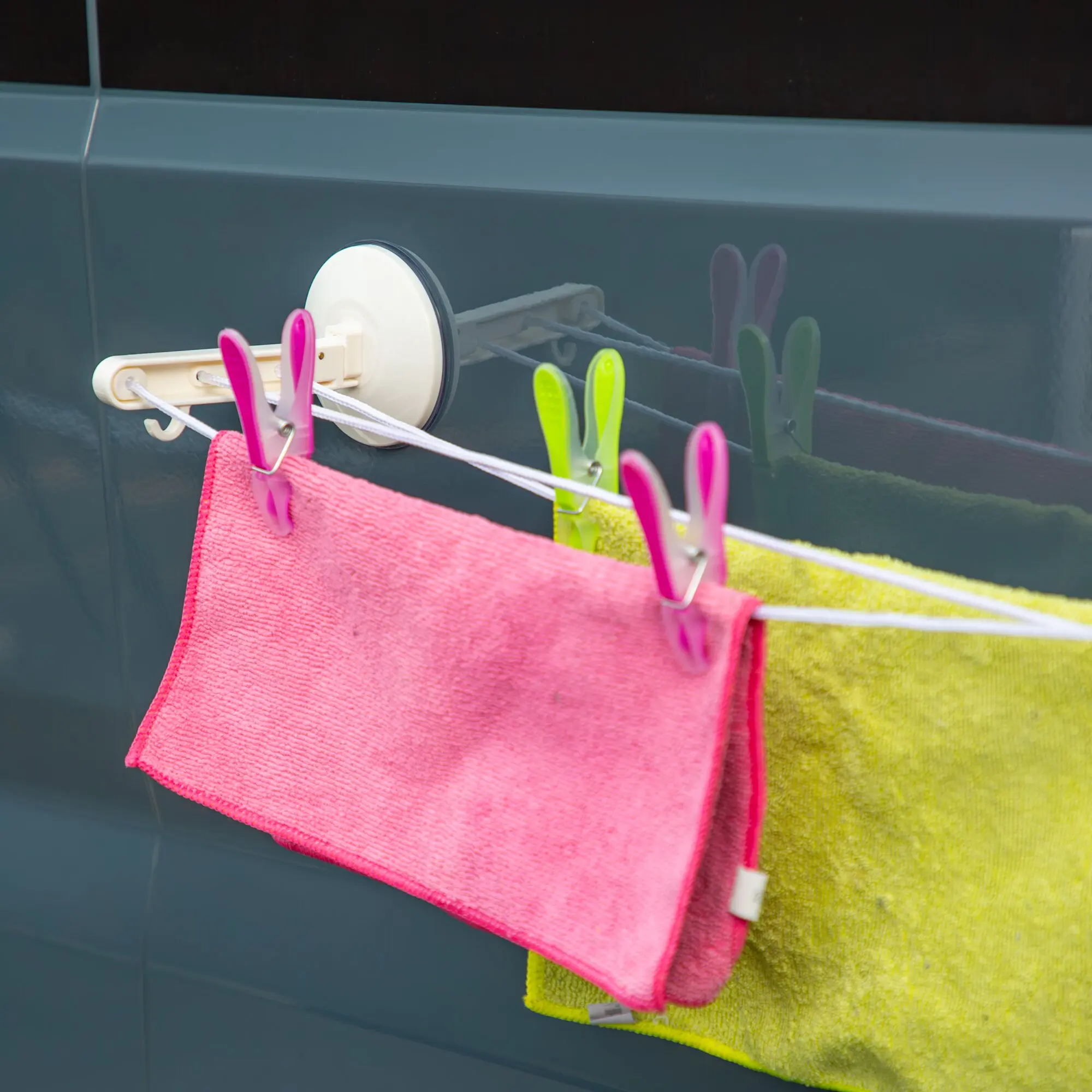 Nahaufnahme eines Wäscheleinen-Systems mit weißem Spulenteil, bunten Wäscheklammern in Pink und Neon-Grün sowie pinkem und limegrünem Handtuch an der Leine
