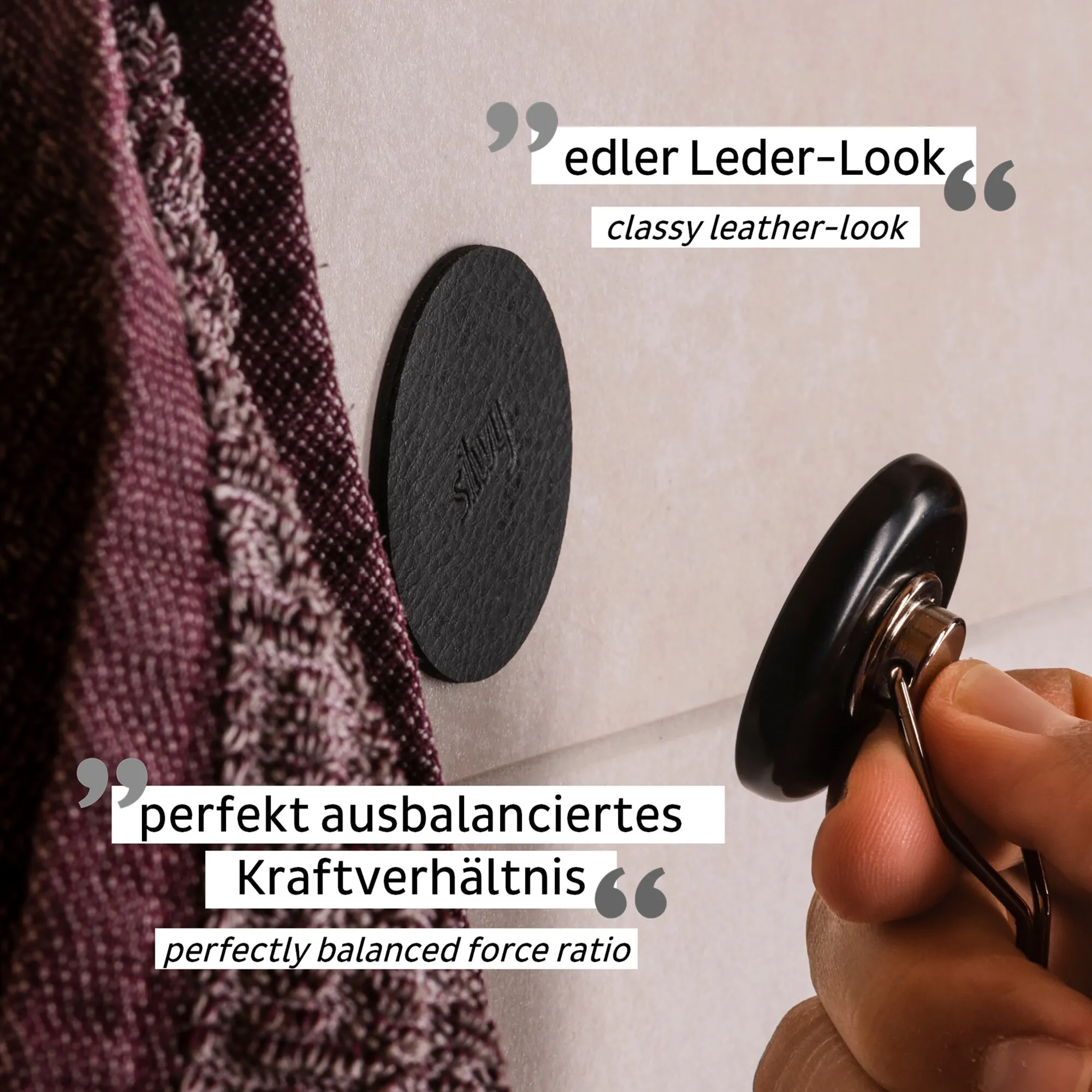 Detailaufnahme des schwarzen Magnet-Hakens THE ONE im Leder-Look: Eine Hand hält den Haken neben der ovalen Wandhalterung, links hängt Stoff