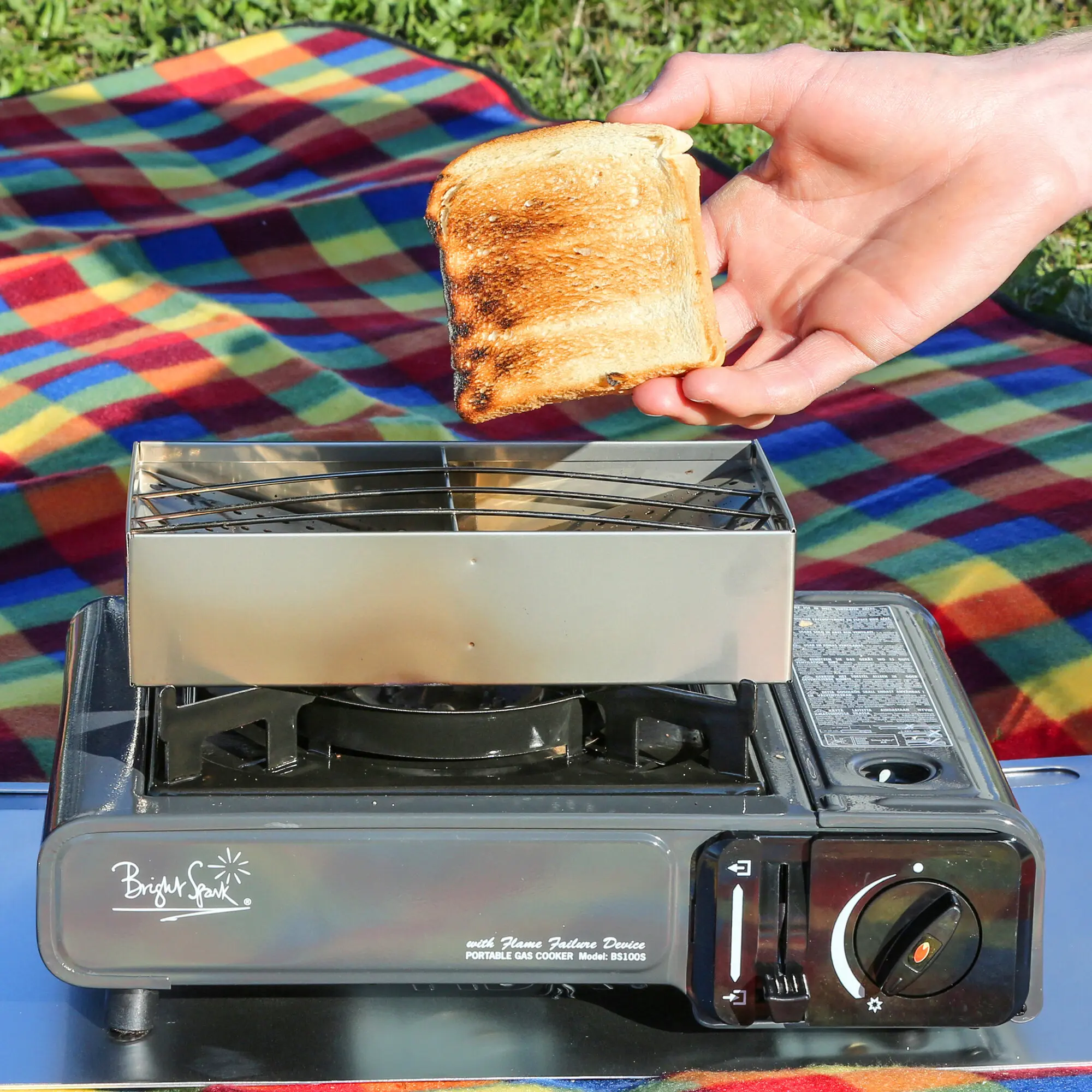 Eine Hand hält eine braun getoastete Brotscheibe über einem kompakten tragbaren Gasgrill, davor eine farbenfrohe Picknickdecke im Freien