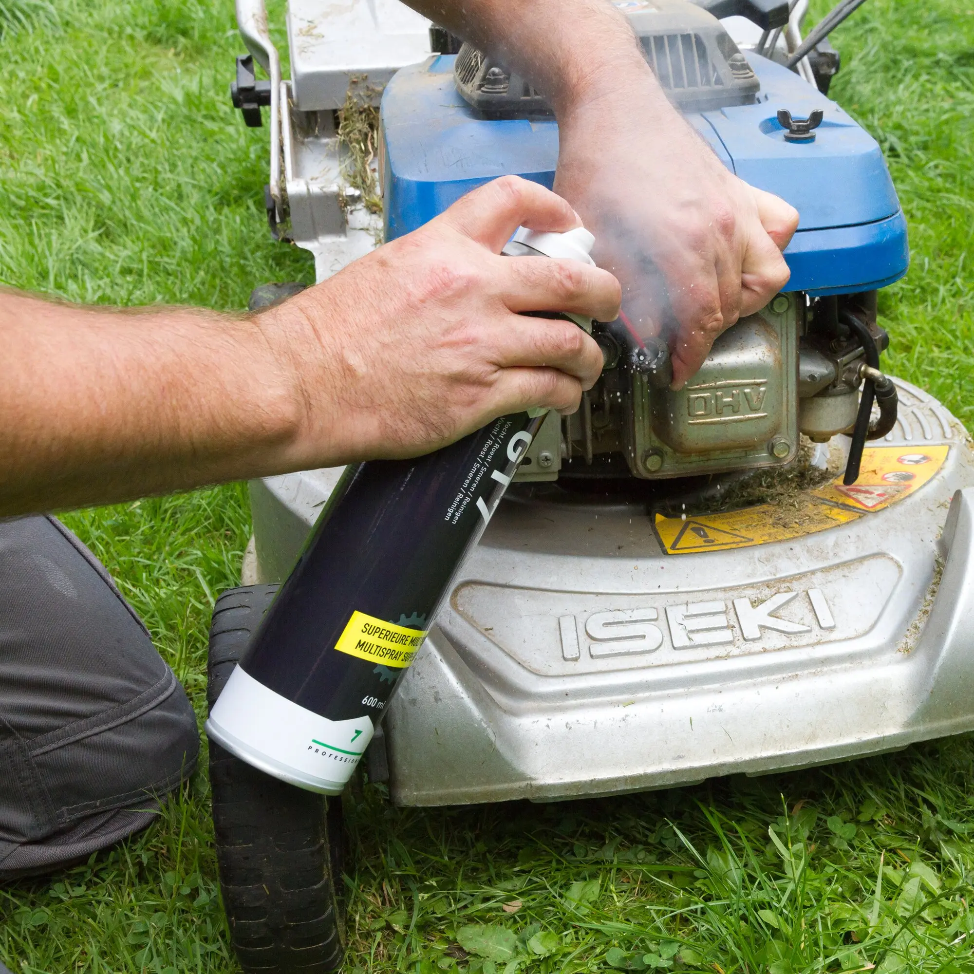 Eine Person verwendet das GT7 Multifunktionsspray auf dem blauen Motor eines Rasenmähers, Gras umgibt die Maschine