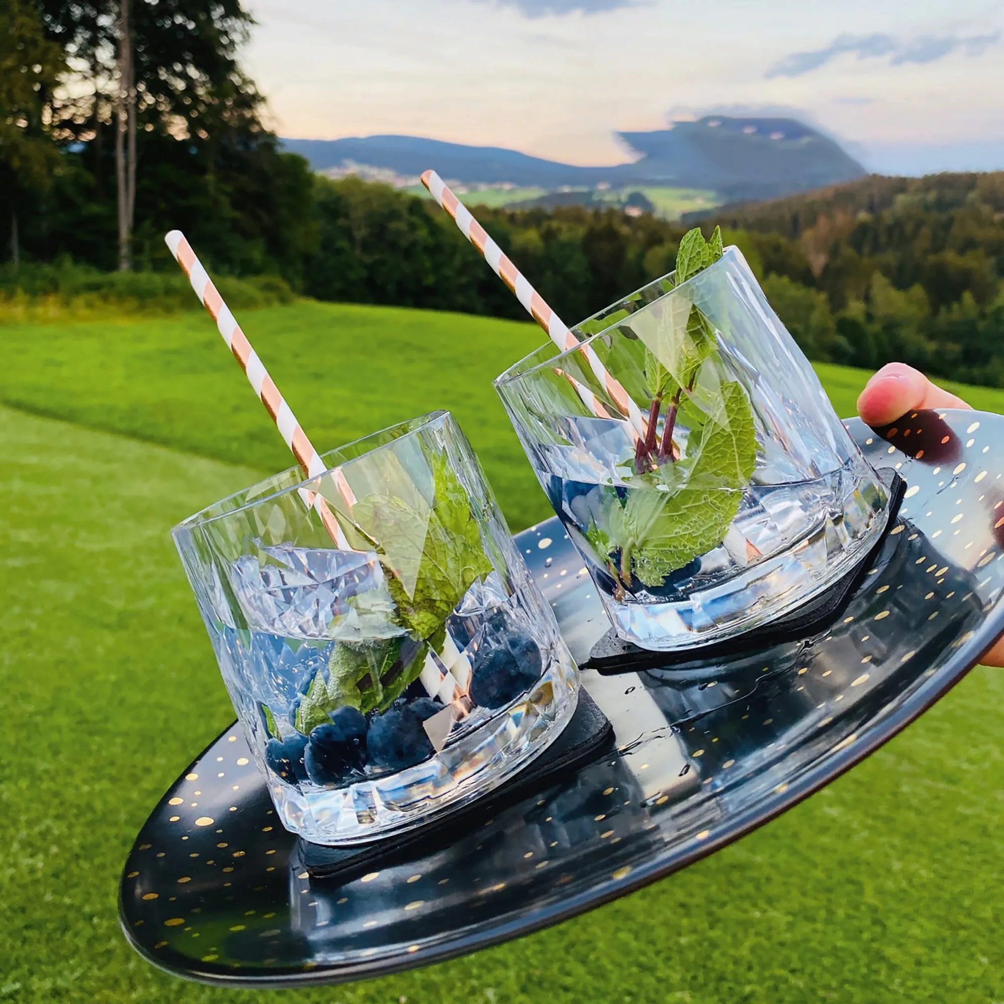 Zwei transparente Magnet-Kunststoffgläser für Whisky mit Eis, Minze, Blaubeeren und Strohhalmen auf einem schwarzen Tablett vor Berglandschaft