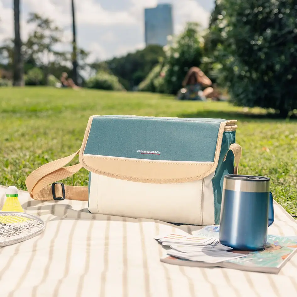 Kühltasche in blau-türkis mit beigem Rand liegt auf Picknickdecke im Park; daneben blauer Thermobecher und Spielkarten, Hintergrund unscharf