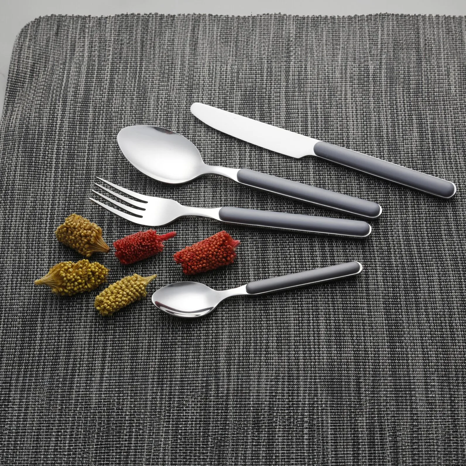 Graues Besteckset Delice mit Messer, Gabel, großem Löffel und kleinem Löffel auf grauer Tischunterlage neben Trockenblumen