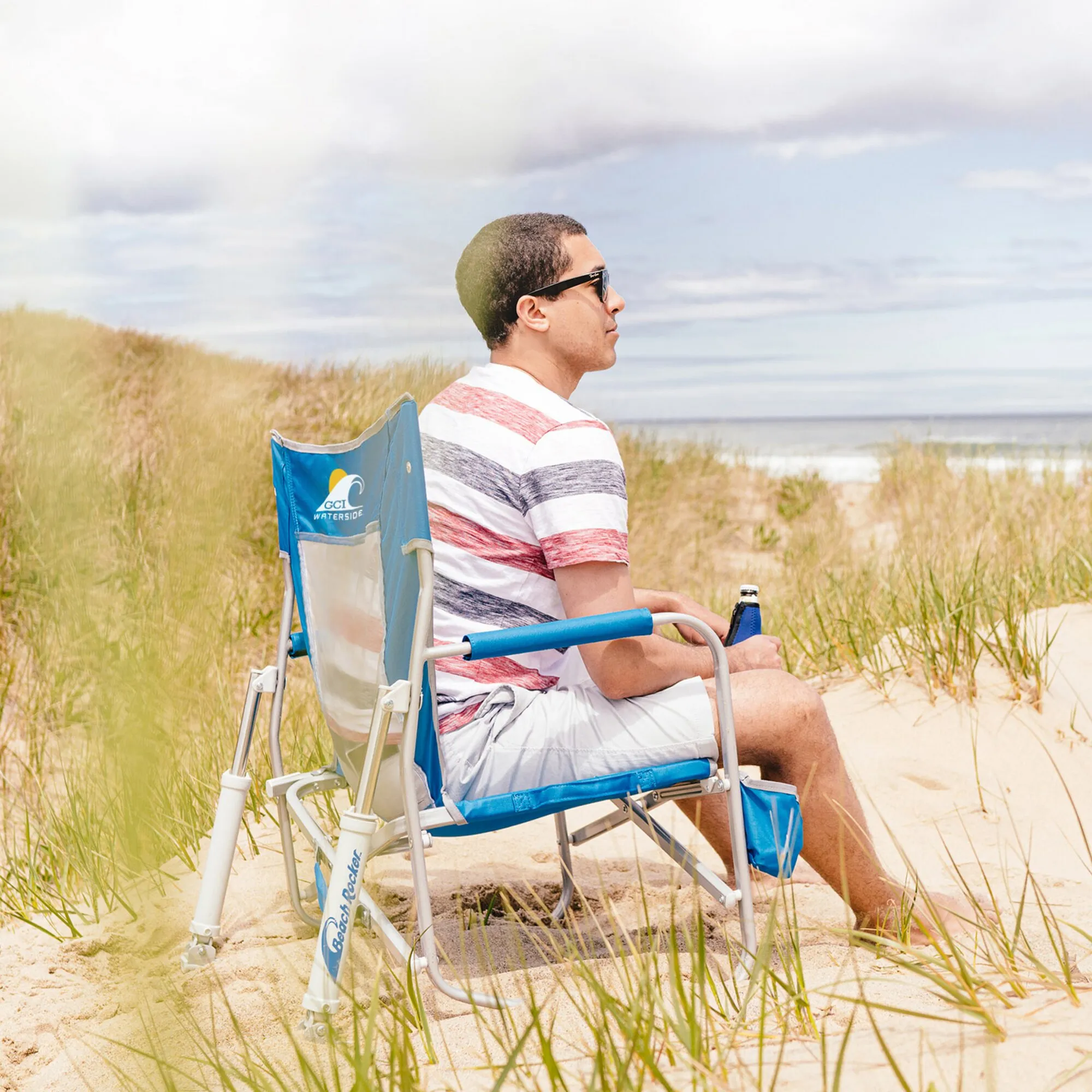 Mann sitzt im blauen Beach Rocker Camping-Schaukelstuhl mit Getränkehalter am Sandstrand zwischen Dünengras
