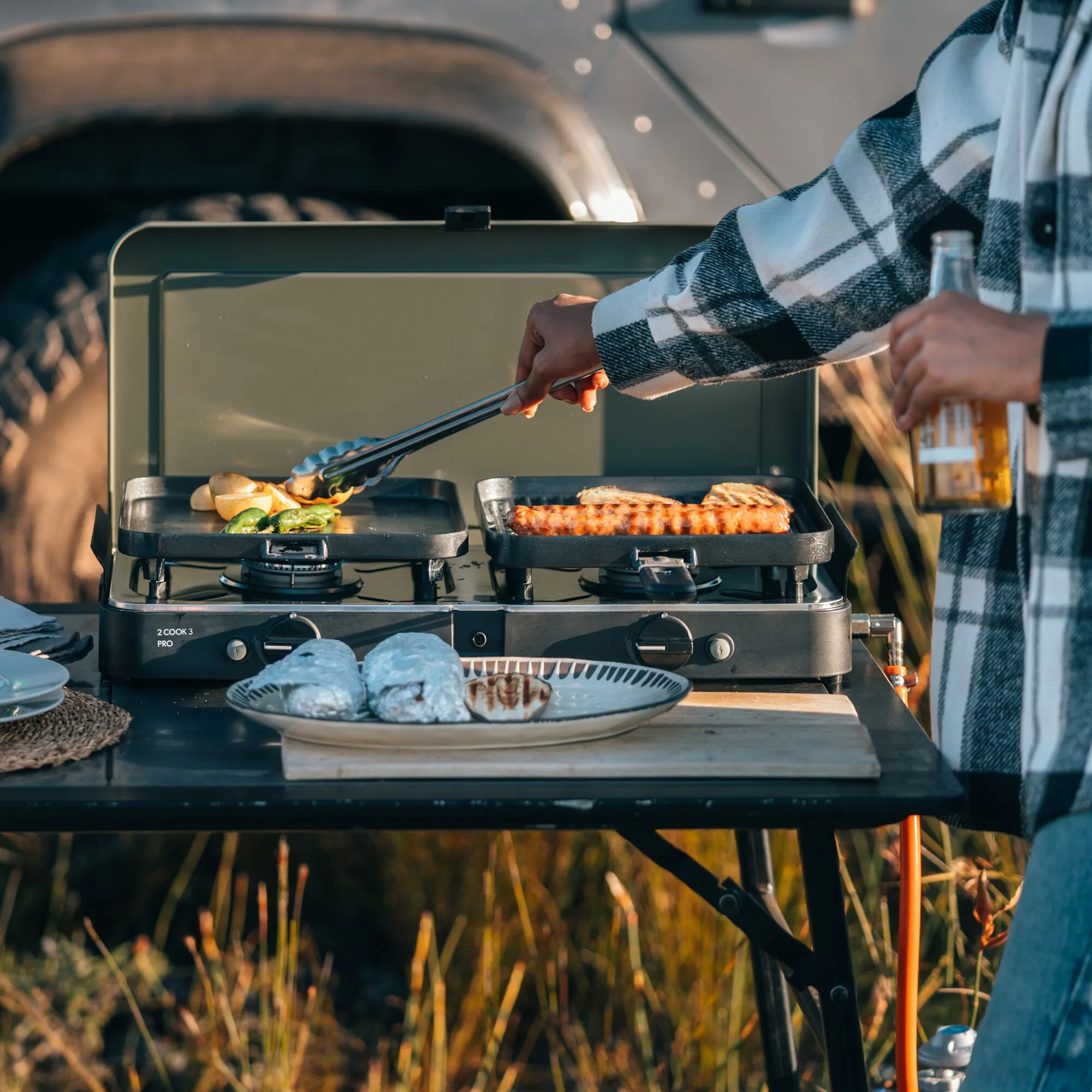 Gaskocher 2 Cook 3 Pro beim Kochen im Freien: Zwei Grillpfannen mit Gemüse und Würstchen vor einem Campingbus