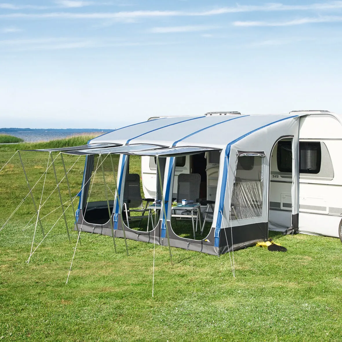 Weiß-blaues Teilzelt Space Air HQ, aufgebaut an einem Wohnwagen mit transparenten Fenstern, Stühlen und Tisch auf einer Wiese am Meer