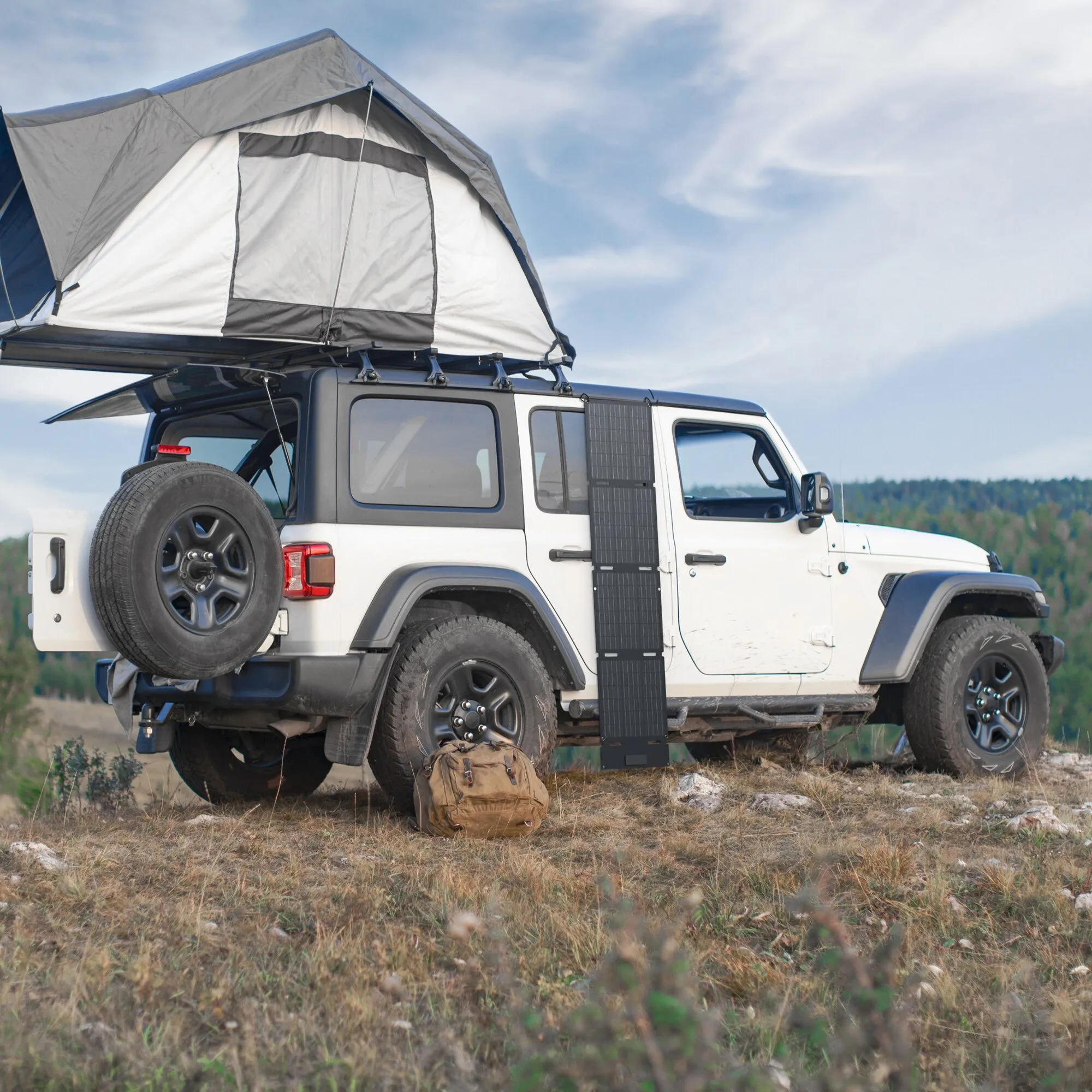 Weißer Geländewagen mit aufklappbarem Dachzelt, seitlich montiertes faltbares Solarmodul, Ersatzrad am Heck, braune Tasche im Vordergrund