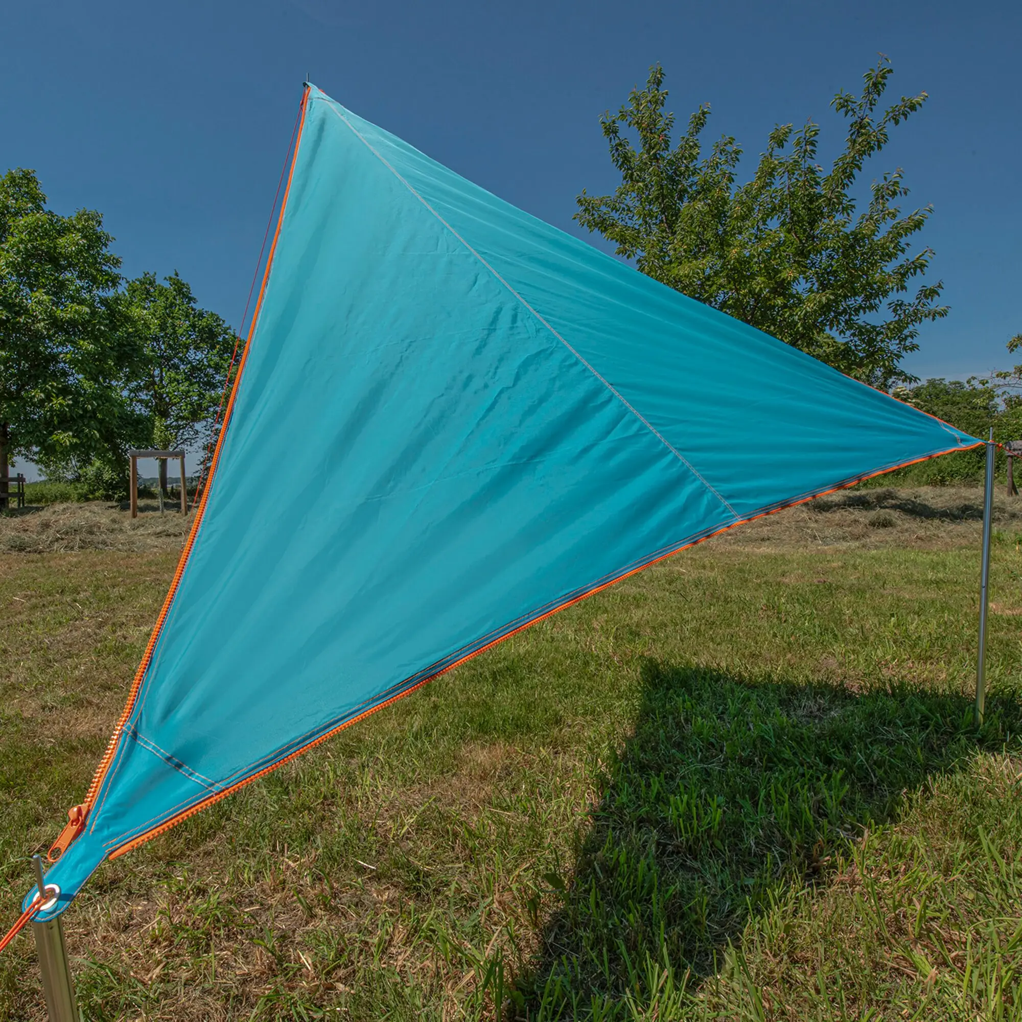 Türkises dreieckiges Sonnensegel mit orangefarbenem Keder und sichtbaren Abspannseilen auf Gras, vor Bäumen und blauem Himmel installiert